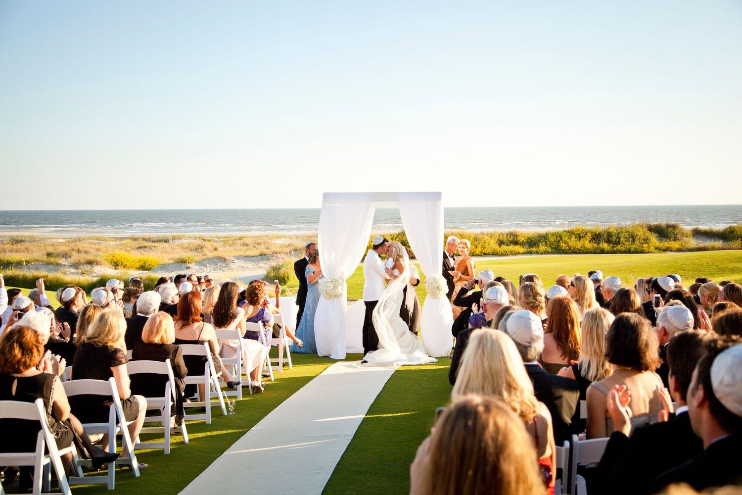 Outdoor wedding ceremony overlooking the ocean with white chairs and aisle runner under clear blue skies.