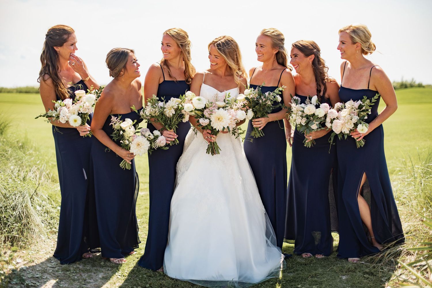 A row of bridesmaids in navy blue dresses stand with a bride in a white gown holding floral bouquets outdoors.
