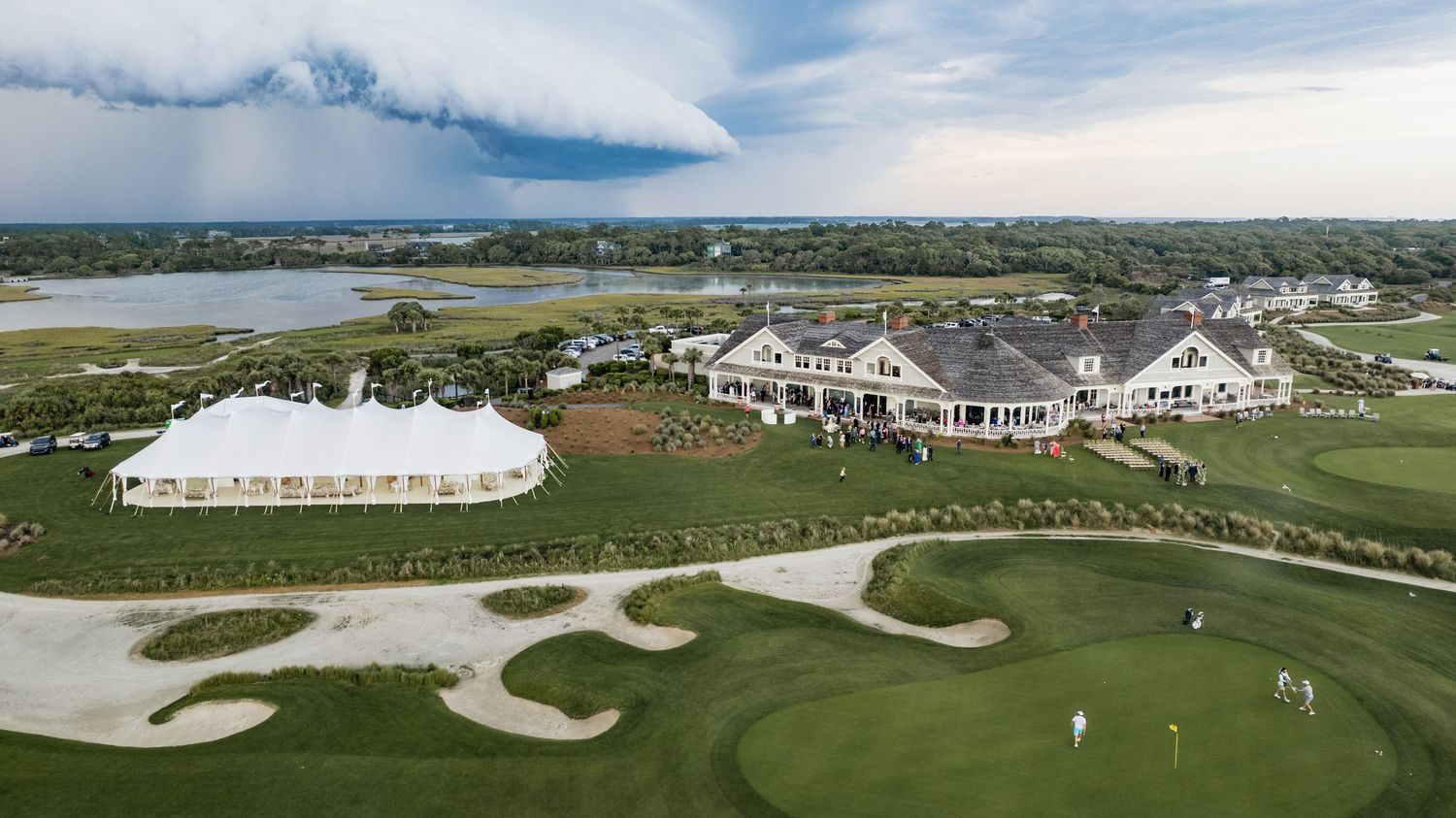 Aerial panoramic view of a golf course with white buildings and sand traps under a cloudy blue sky.