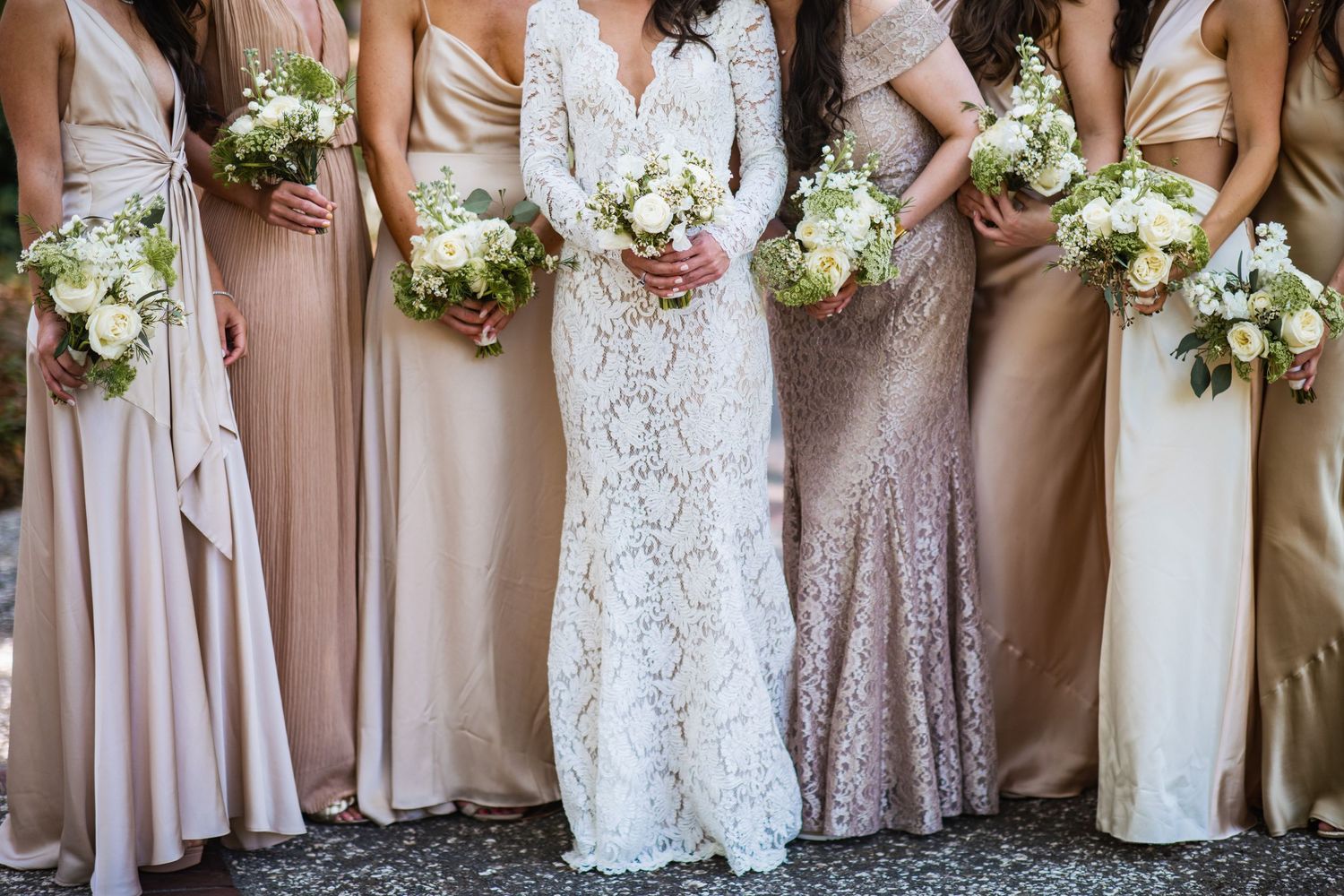 Elegant bridal party in blush pink and neutral dresses holding white floral bouquets lined up for formal photo.