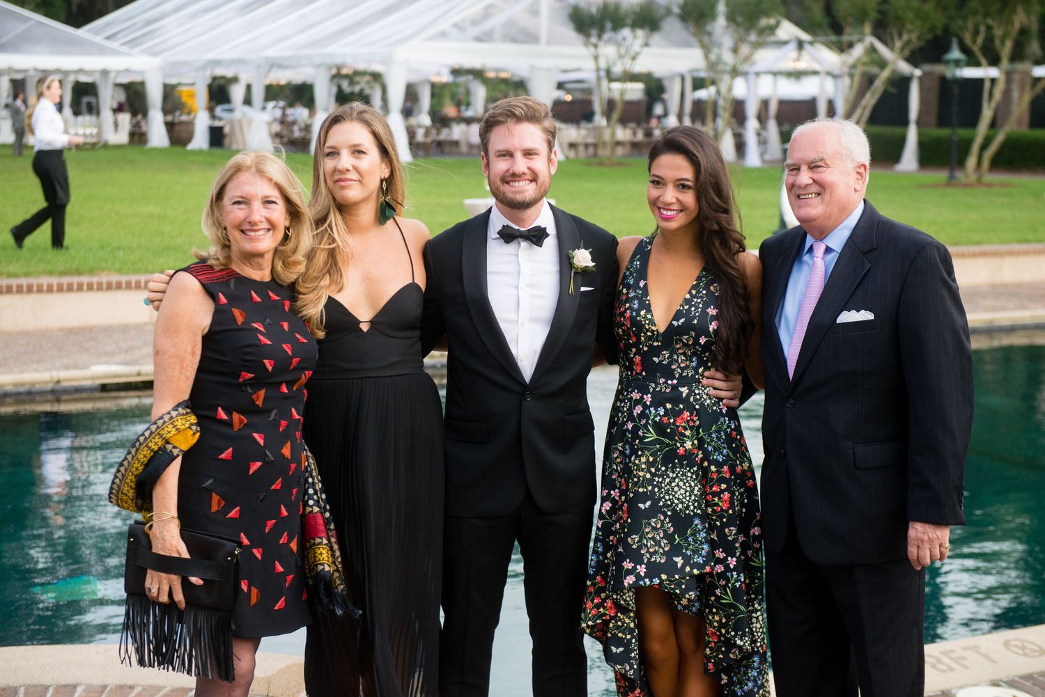 A group of formally dressed people pose together at an outdoor event near a swimming pool.