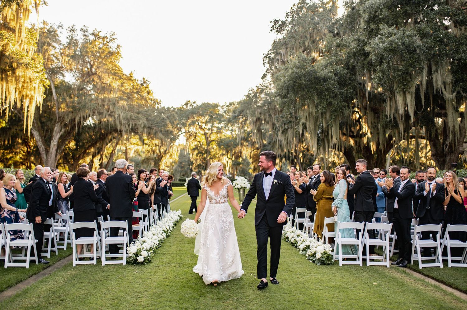 A scenic outdoor wedding ceremony under moss-draped oak trees with white chairs and golden sunlight filtering through.