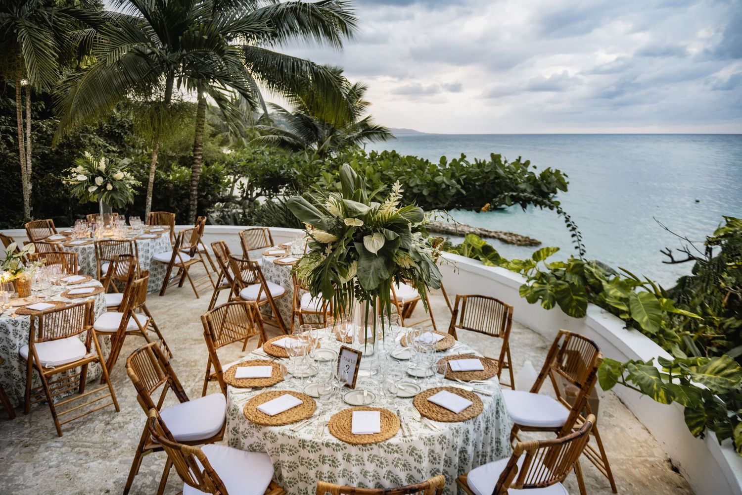 Elegant beach wedding reception setup with wooden chairs and tables decorated with white linens and palm trees overhead.