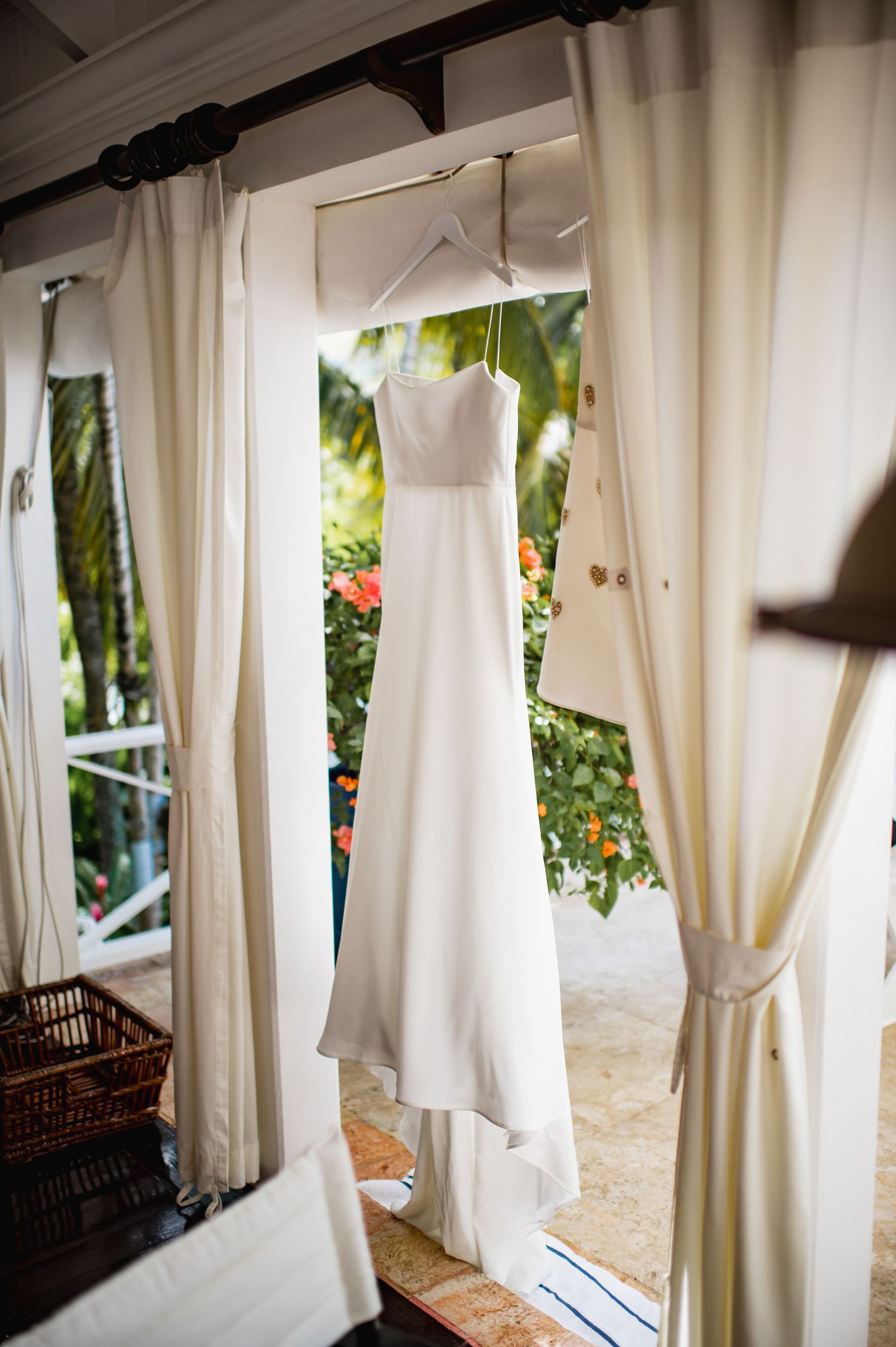 An elegant white wedding dress hangs by a window with flowing curtains and tropical garden views in the background.