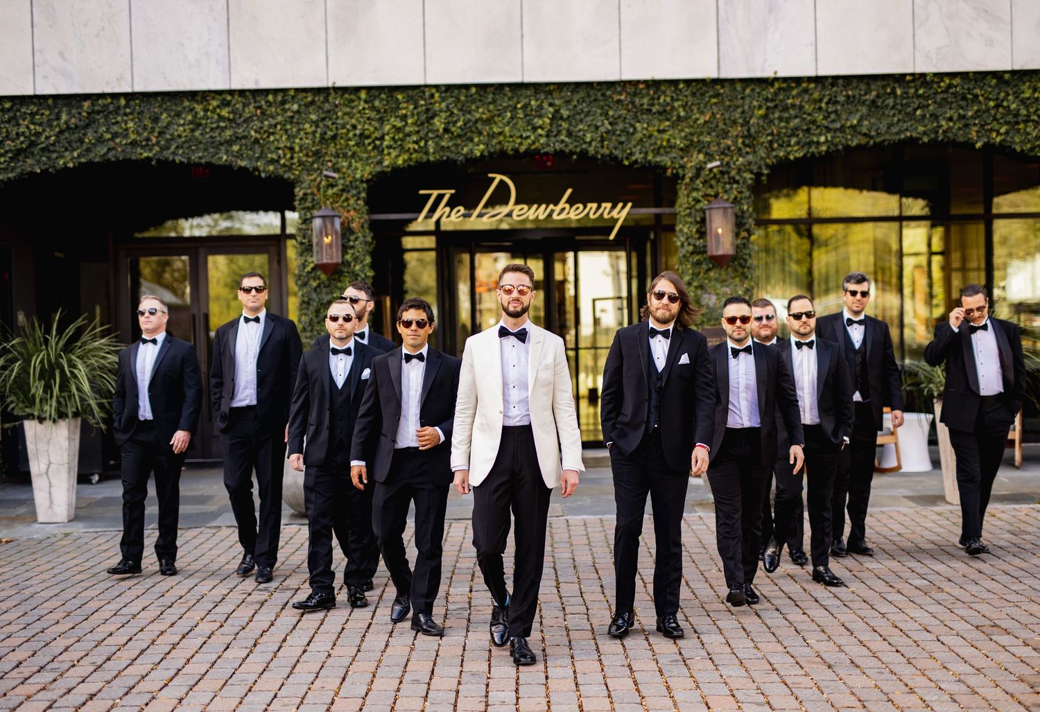 Wedding party members in black tuxedos stand in a line formation outdoors for a formal group photo.