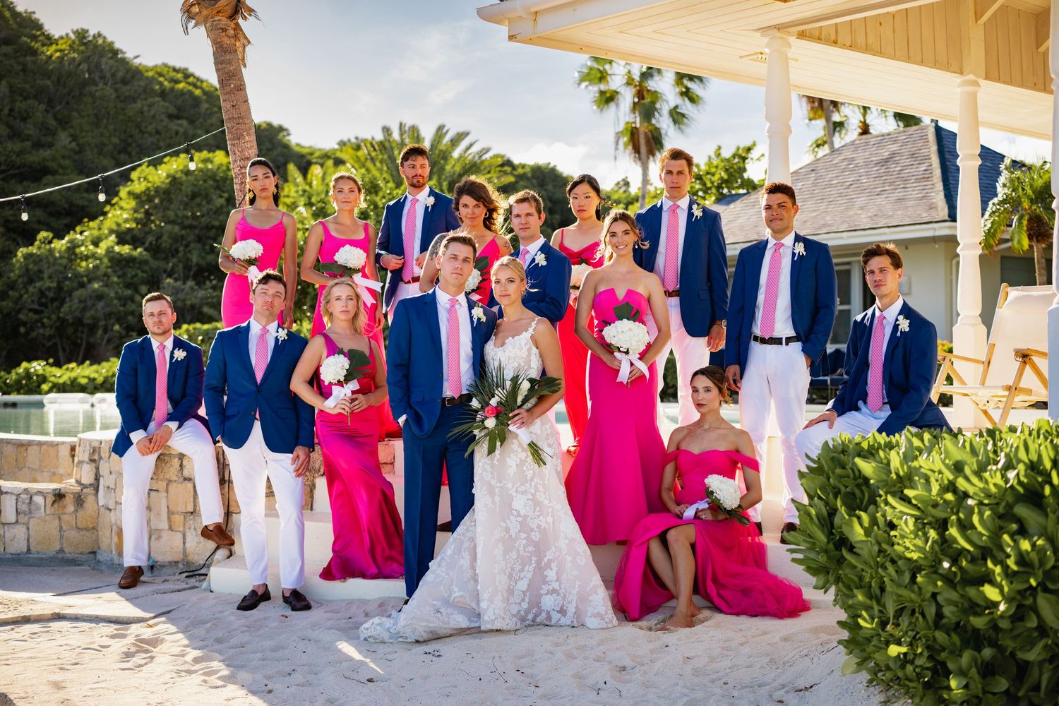 Wedding party in navy blue suits and bright pink dresses pose together at a tropical beach venue.