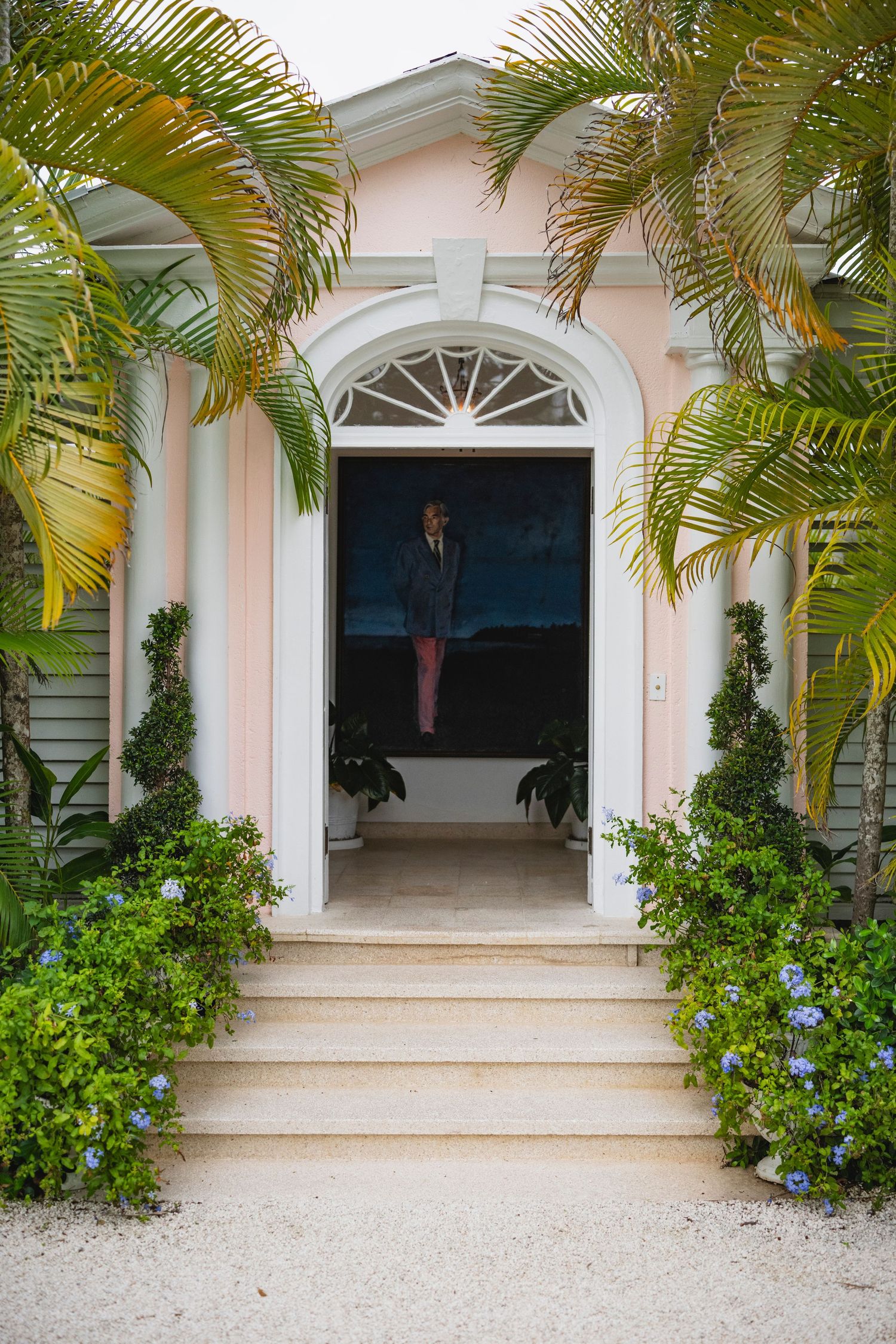 Elegant entryway with pink walls, white arched doorframe, and palm trees framing stone steps to a tropical home.