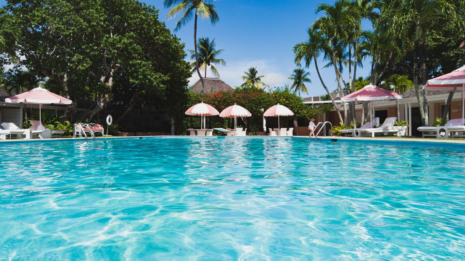 Crystal clear turquoise swimming pool surrounded by palm trees and white umbrellas at a luxury tropical resort.