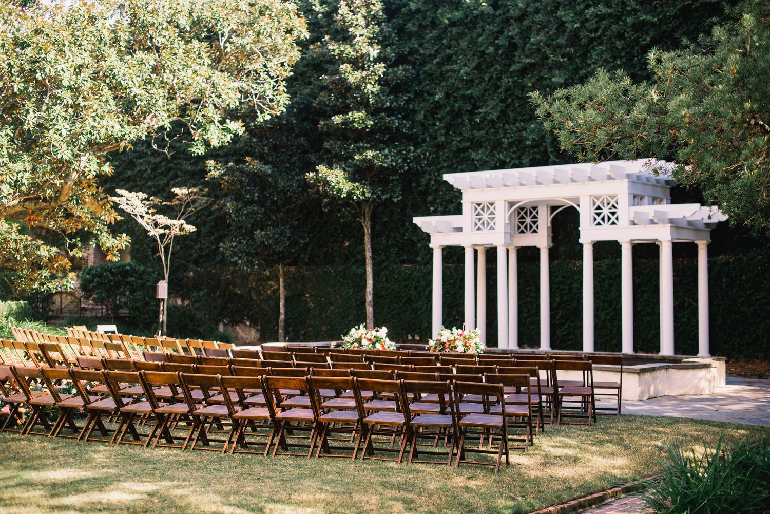 Elegant white garden pergola with columns set up for an outdoor wedding ceremony with wooden folding chairs on the lawn.