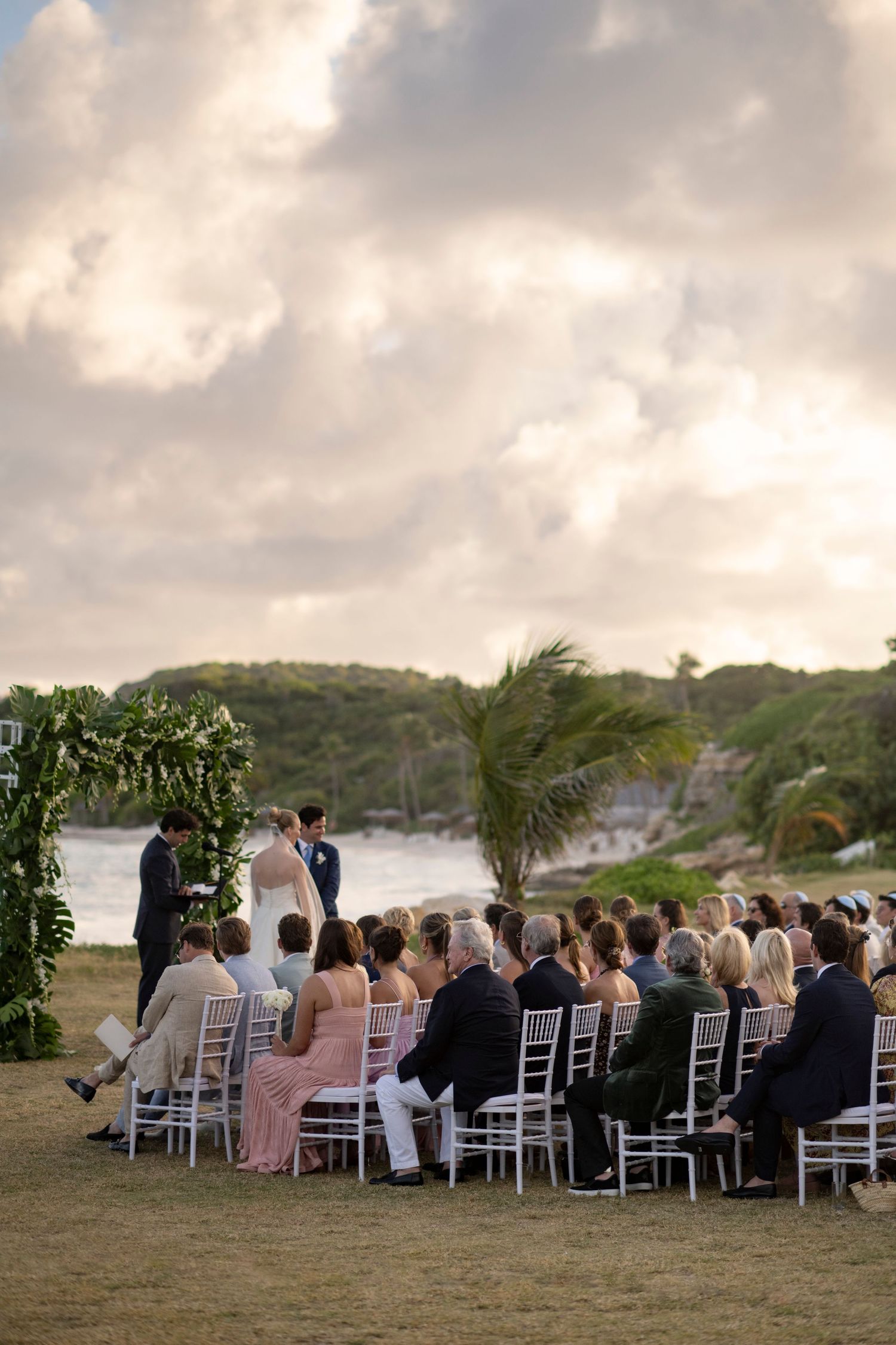 Wedding ceremony guests seated in white chairs overlooking the ocean at sunset.