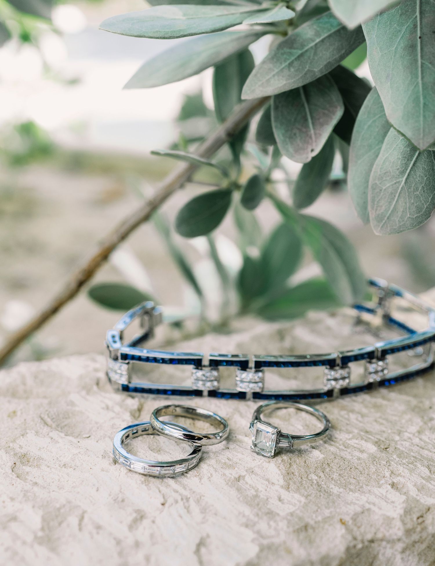 Wedding rings and jewelry arranged on a stone surface with green foliage.
