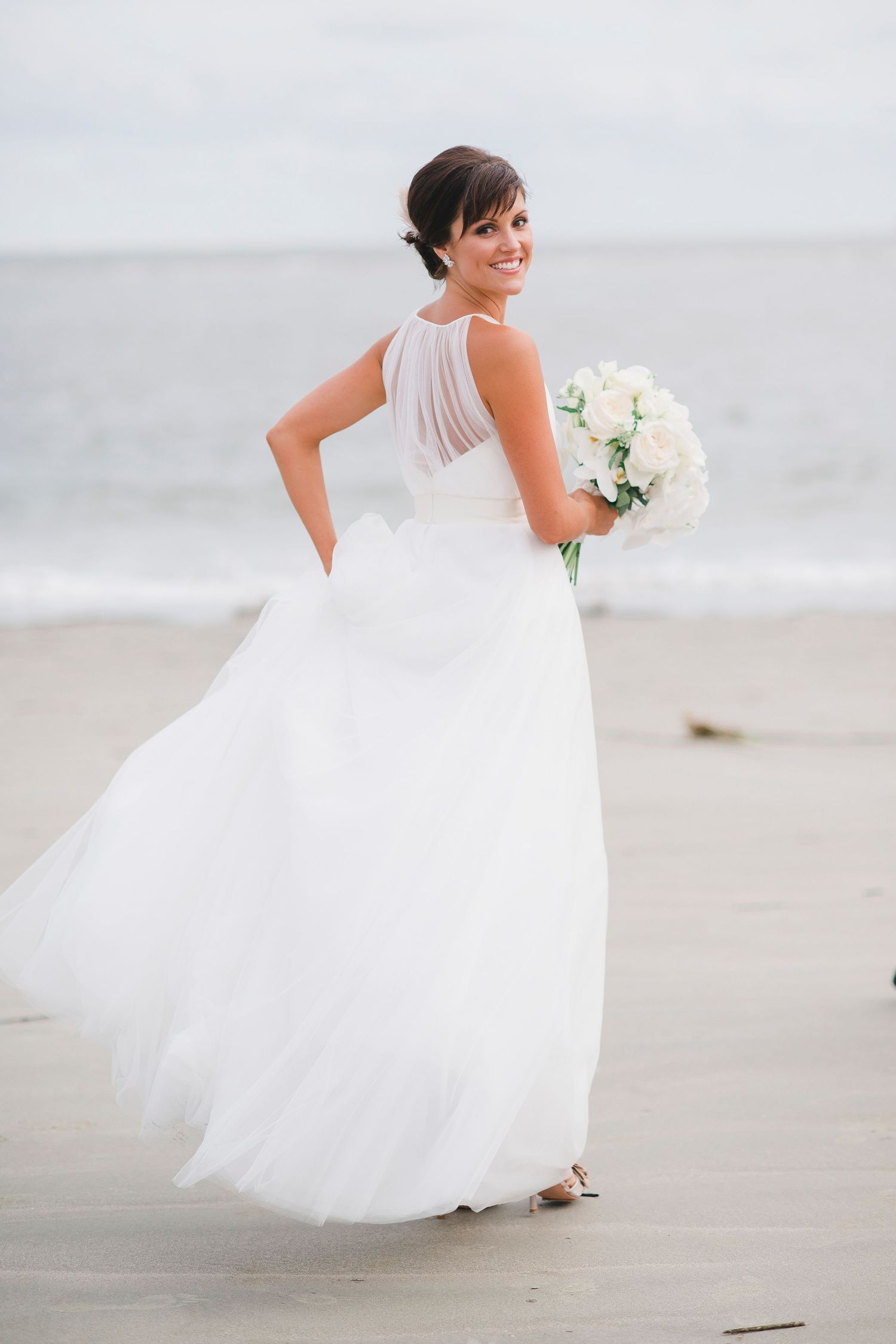 Bride in flowing white dress poses gracefully on sandy beach.