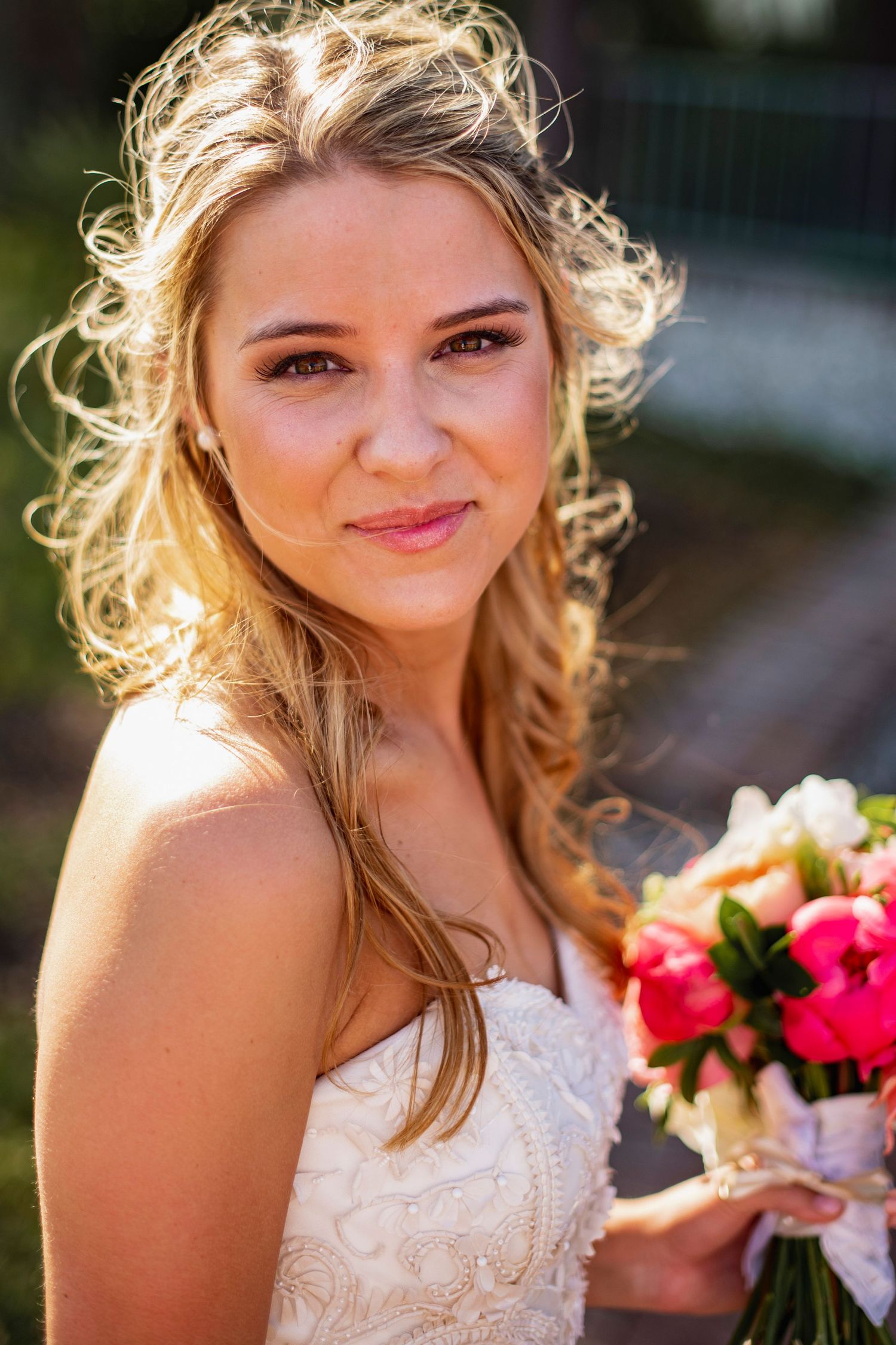 A bride in a white lace dress poses outdoors with sunlit hair and pink bouquet details.