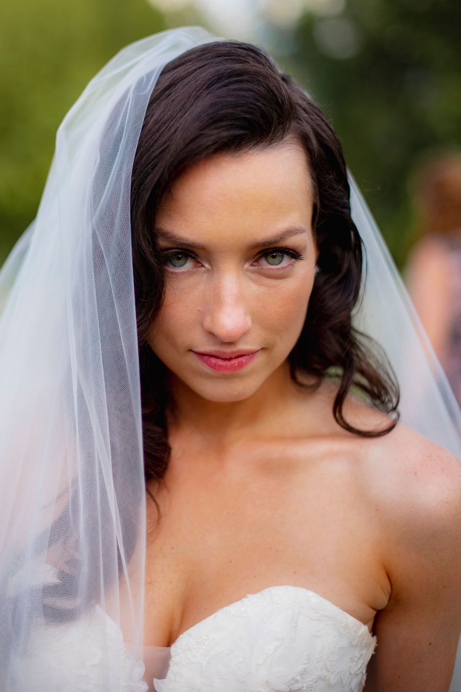 A bride in a strapless white wedding gown and veil smiles while looking down during an outdoor wedding photoshoot.