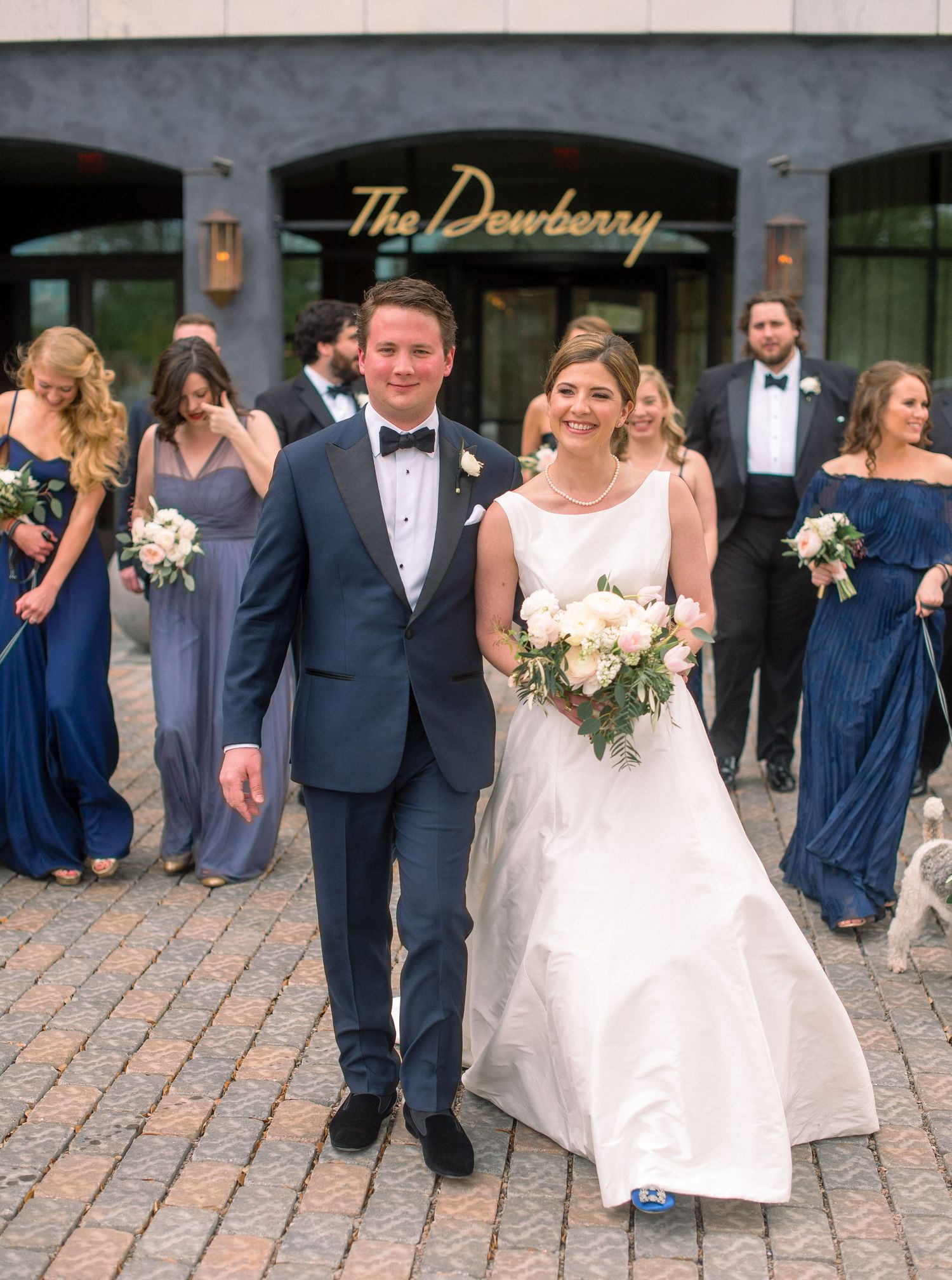 A wedding party poses outside The Dewberry hotel entrance with bridesmaids in navy dresses and white floral bouquets.