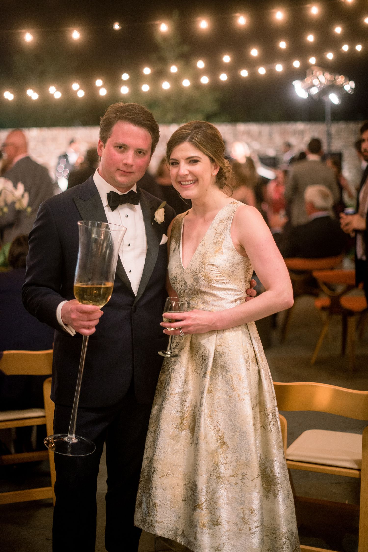 A couple in formal wedding attire holds champagne glasses under romantic string lights at an evening reception.