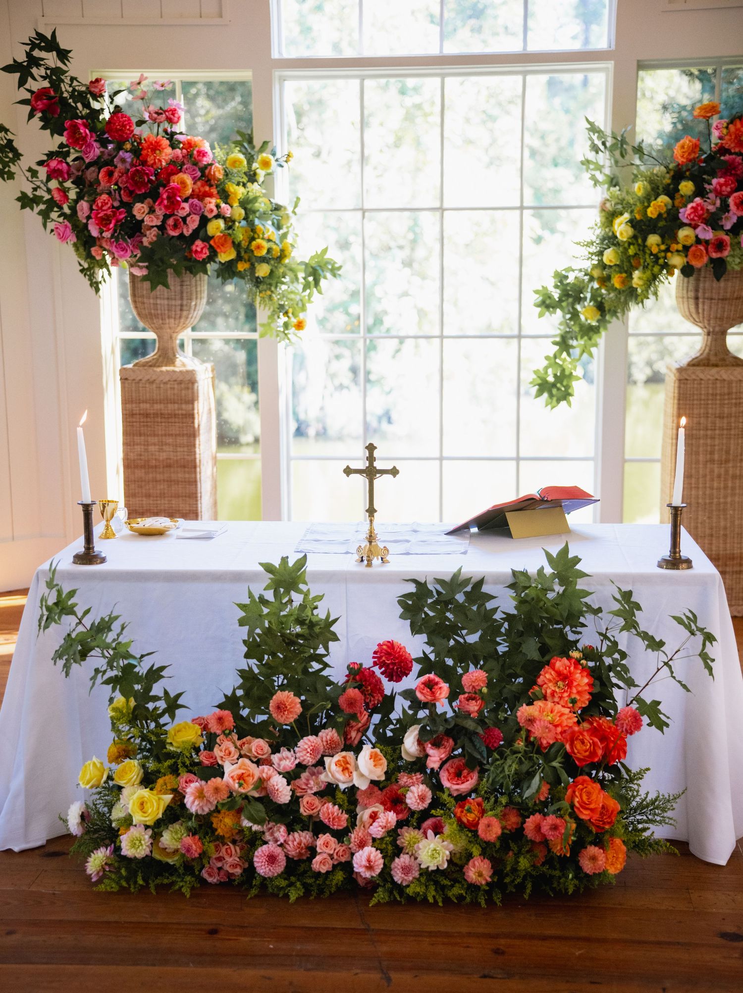 Elegant floral arrangements with vibrant roses and mixed flowers adorn white-clothed tables in a sunlit church or chapel setting.