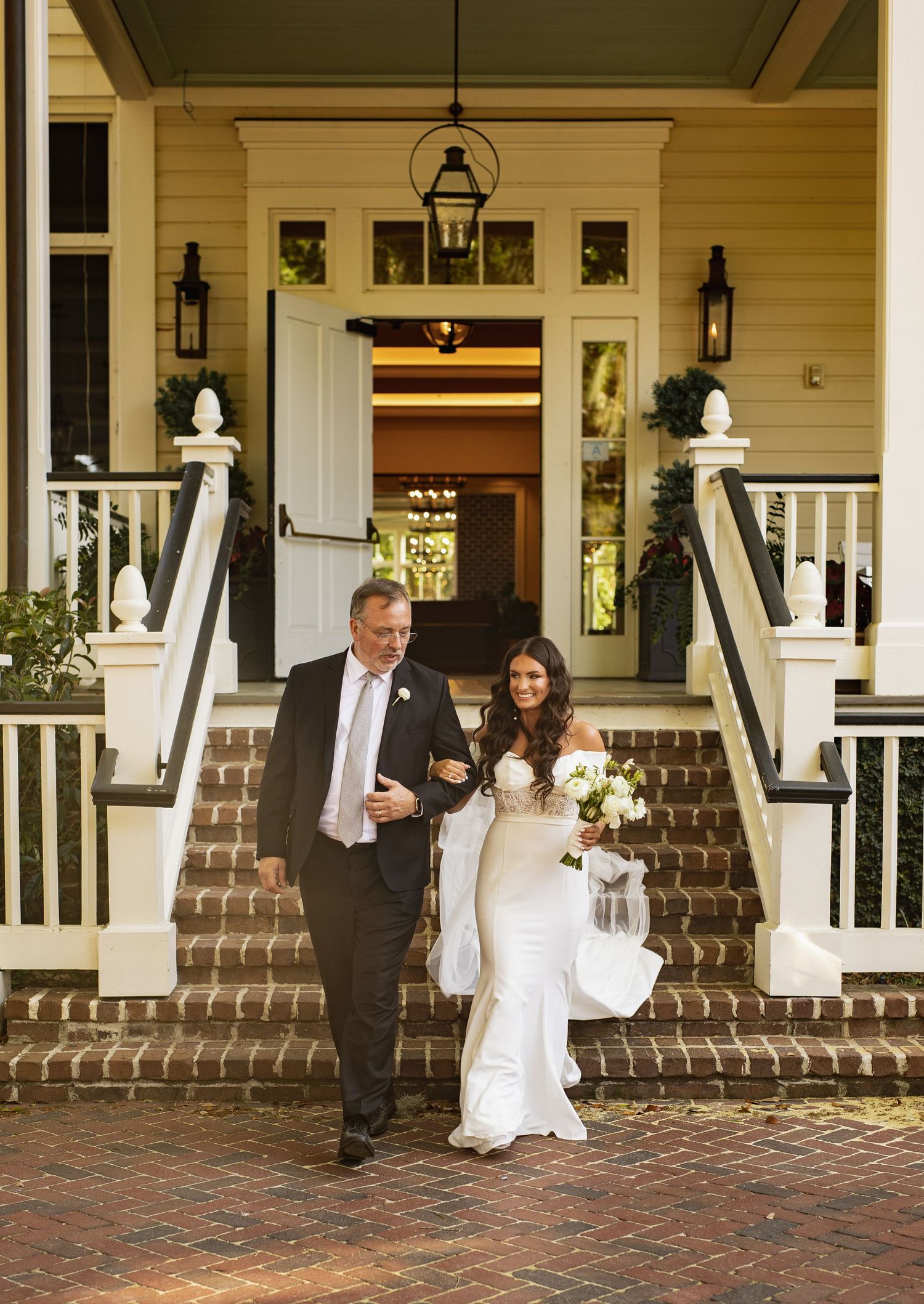 Wedding couple shares playful moments on a charming brick porch with white columns and lanterns at a southern estate.