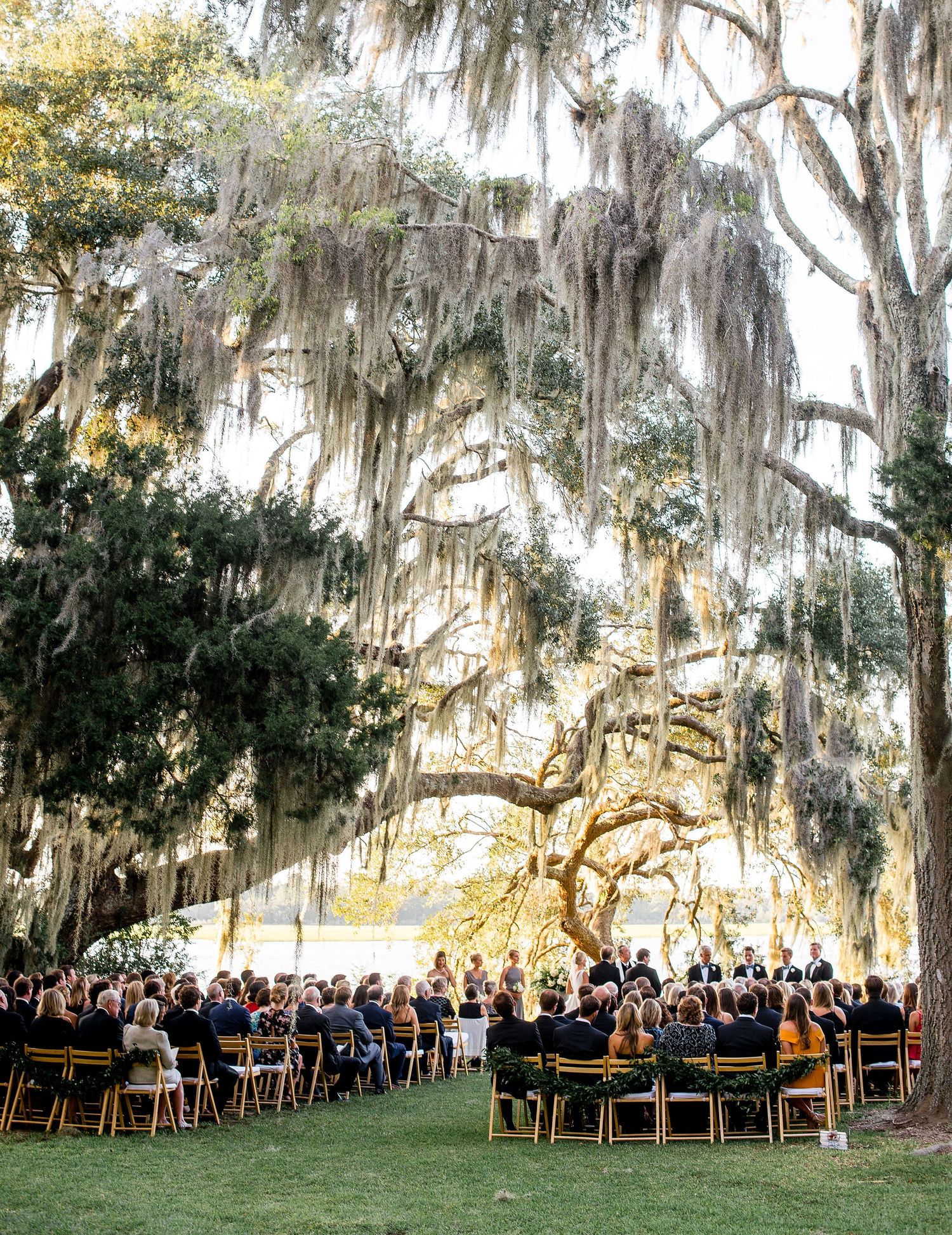 An outdoor wedding ceremony under majestic Spanish moss-draped oak trees with guests seated on wooden chairs.