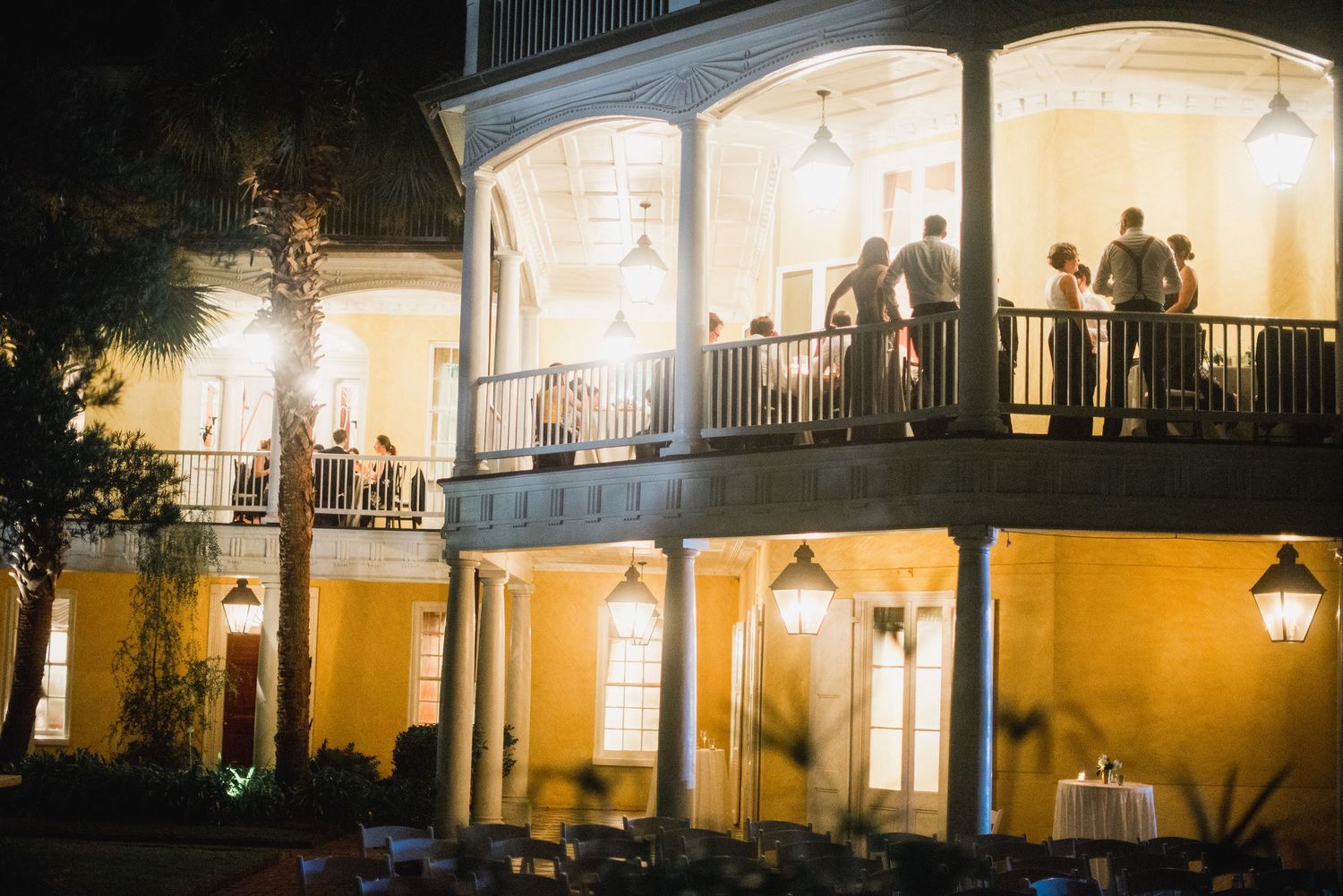 Evening party guests gather on an illuminated yellow mansion's two-story veranda with white columns and warm lighting.
