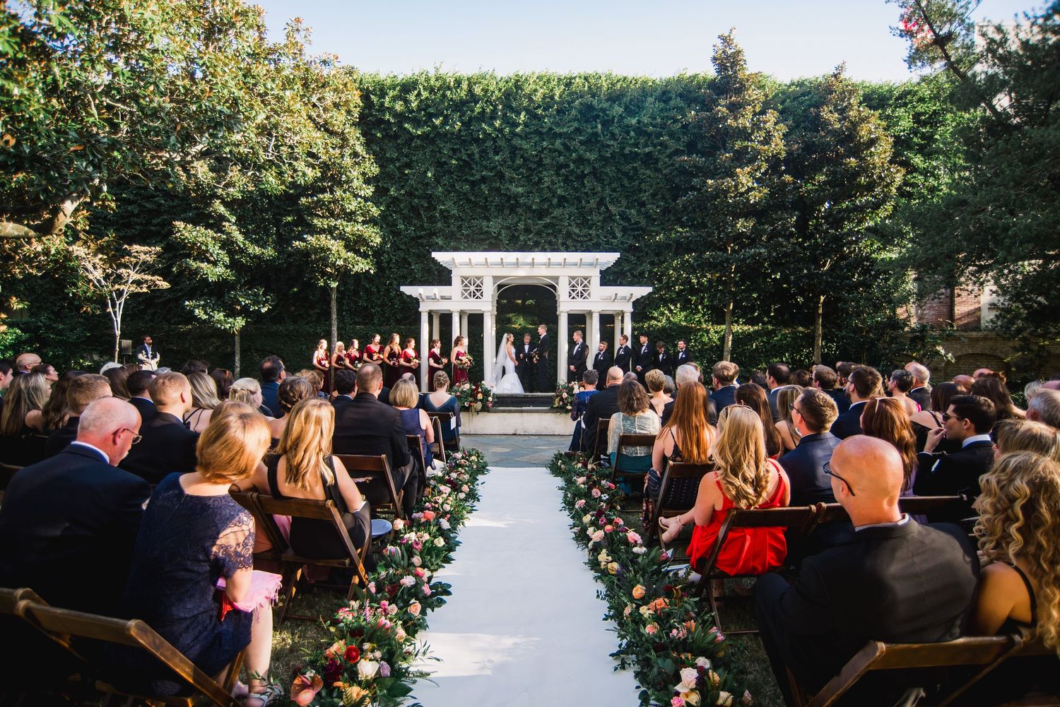 A beautiful outdoor wedding ceremony setup with white chairs and a gazebo surrounded by lush greenery and gardens.