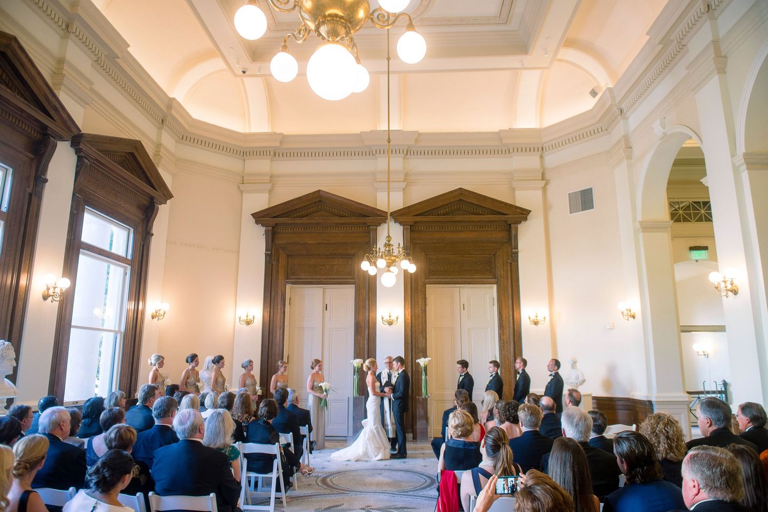 A wedding ceremony takes place in an elegant white room with high ceilings and ornate chandeliers.