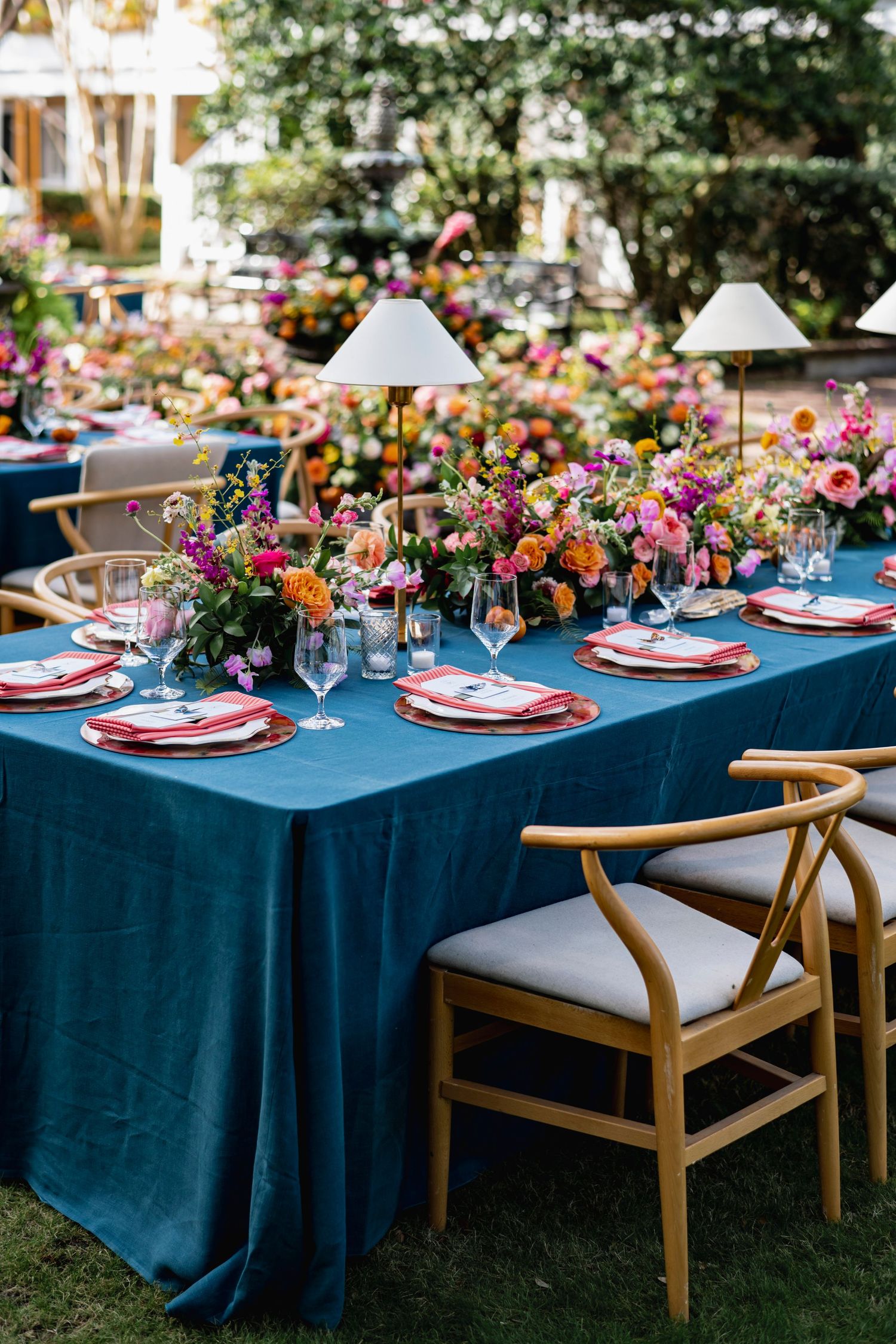 Elegant outdoor dining table with teal blue tablecloth and colorful floral centerpieces set for a garden party.