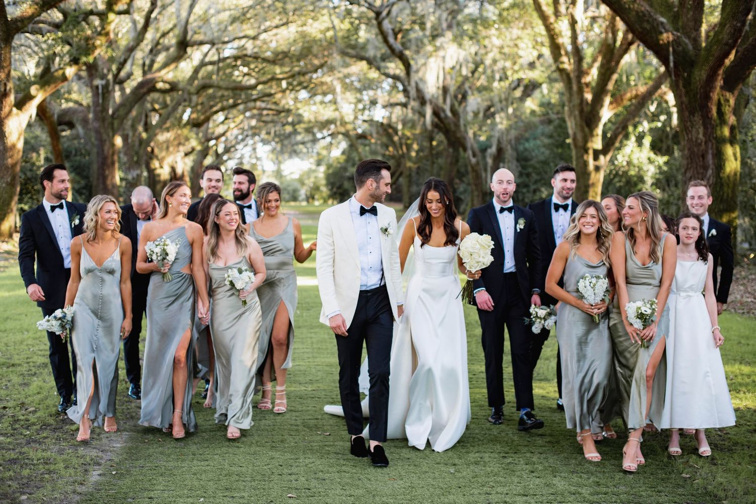 Wedding party in elegant gray dresses and formal attire poses together on a lawn under towering oak trees.