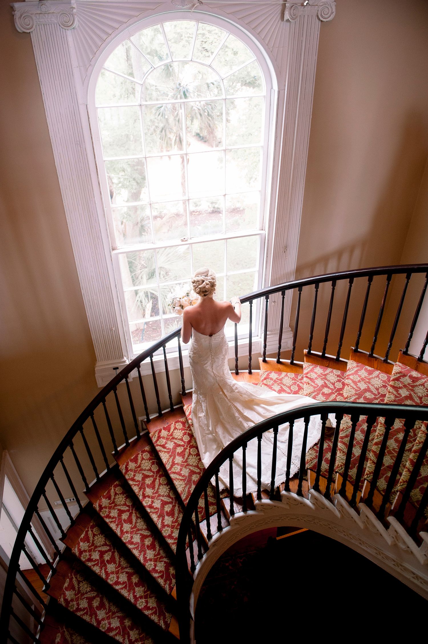 Curved staircase with ornate red and gold carpeting leads up to arched windows in an elegant interior space.