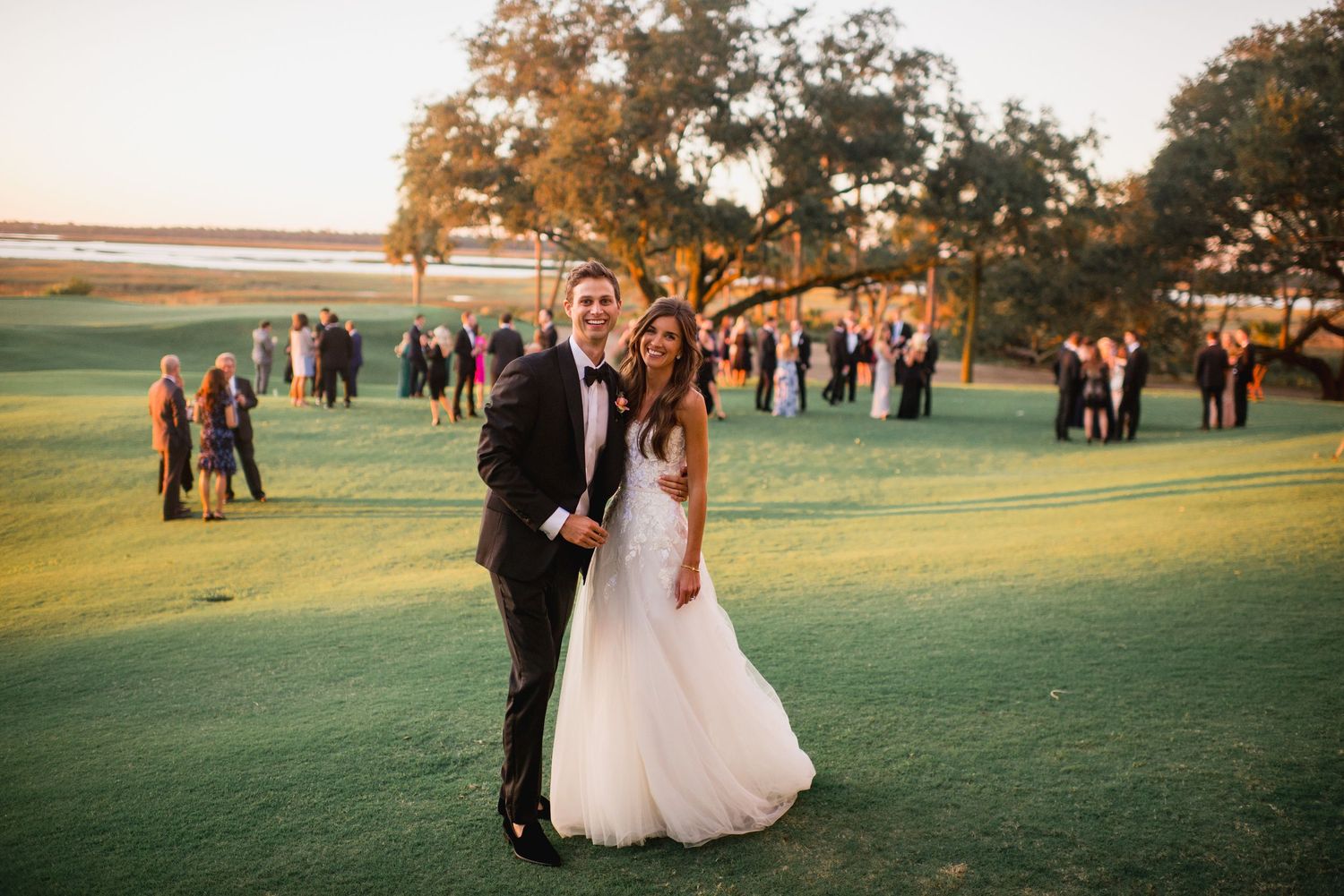 Wedding guests gather on a lush green lawn at sunset with elegant buildings in the background during an outdoor reception.