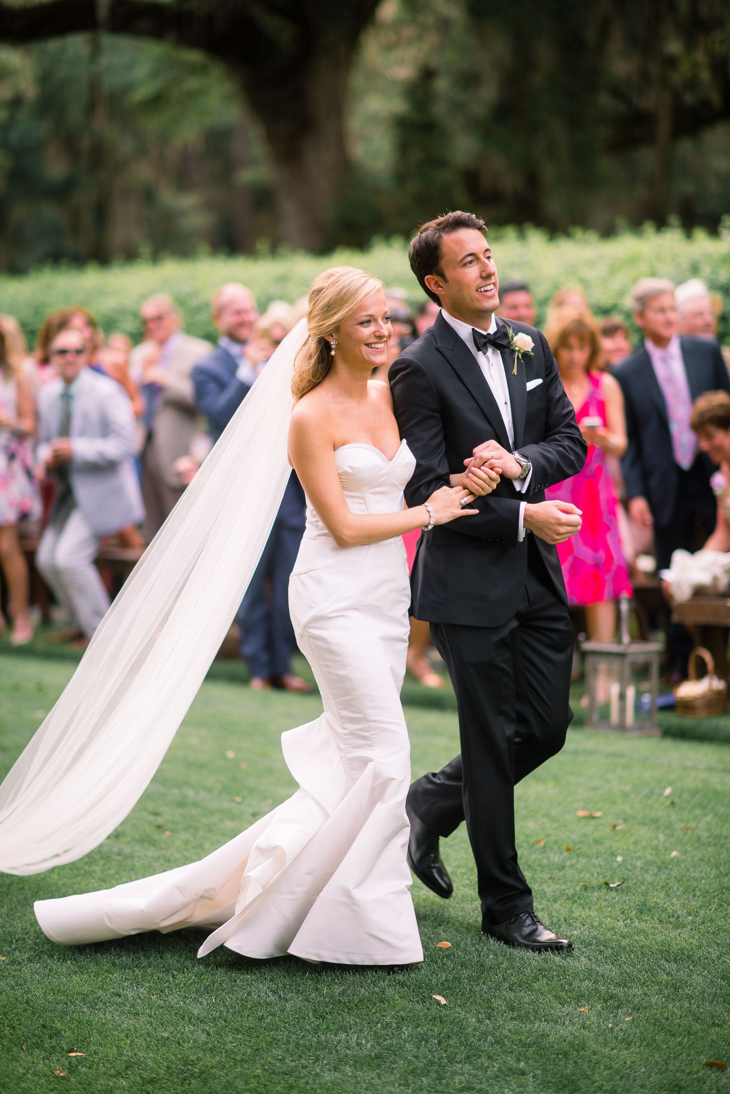 Wedding couple performs a fun dip dance move on green grass lawn with guests watching in background.