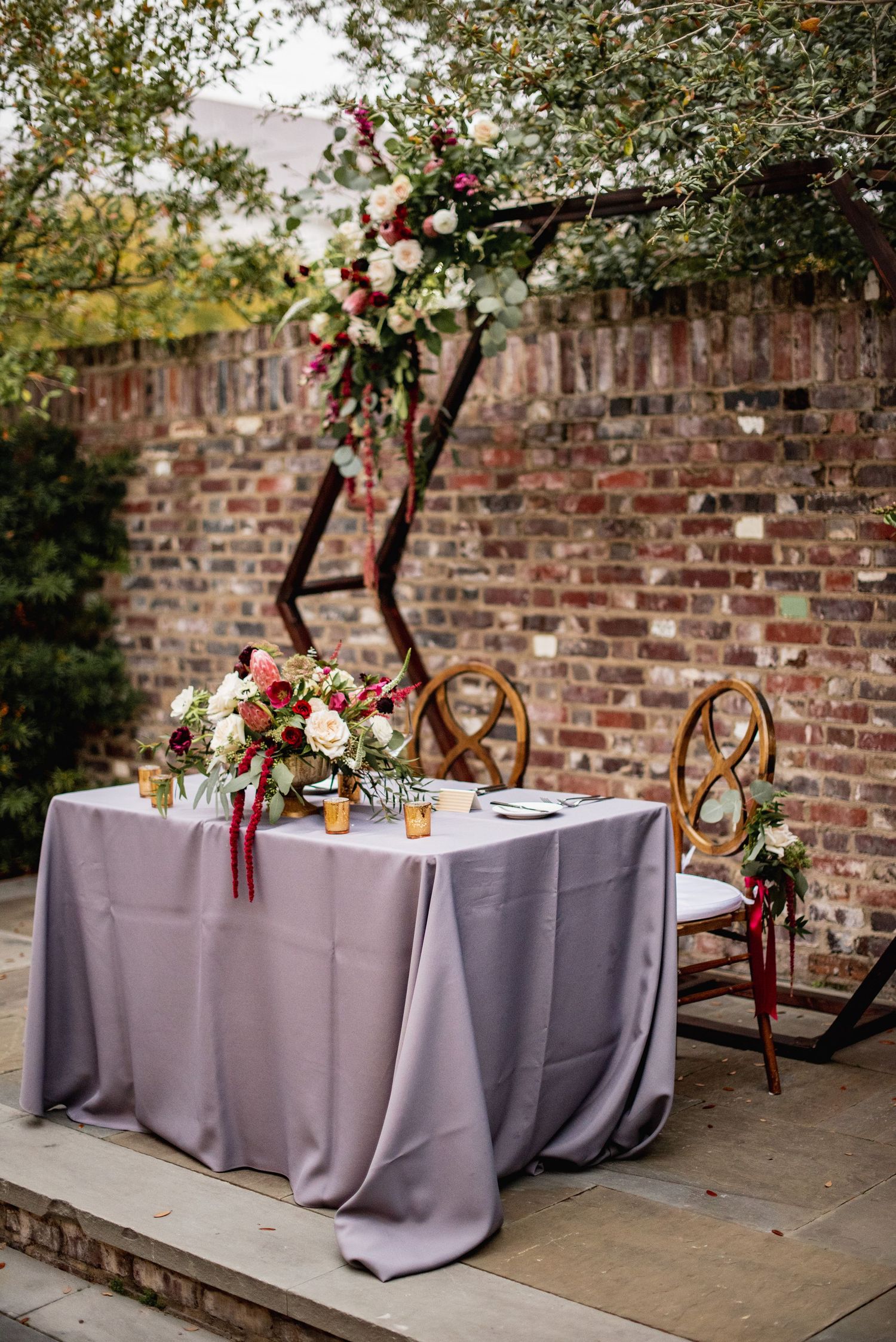Wedding reception tables with lavender tablecloths and floral centerpieces set against rustic brick wall in outdoor venue.