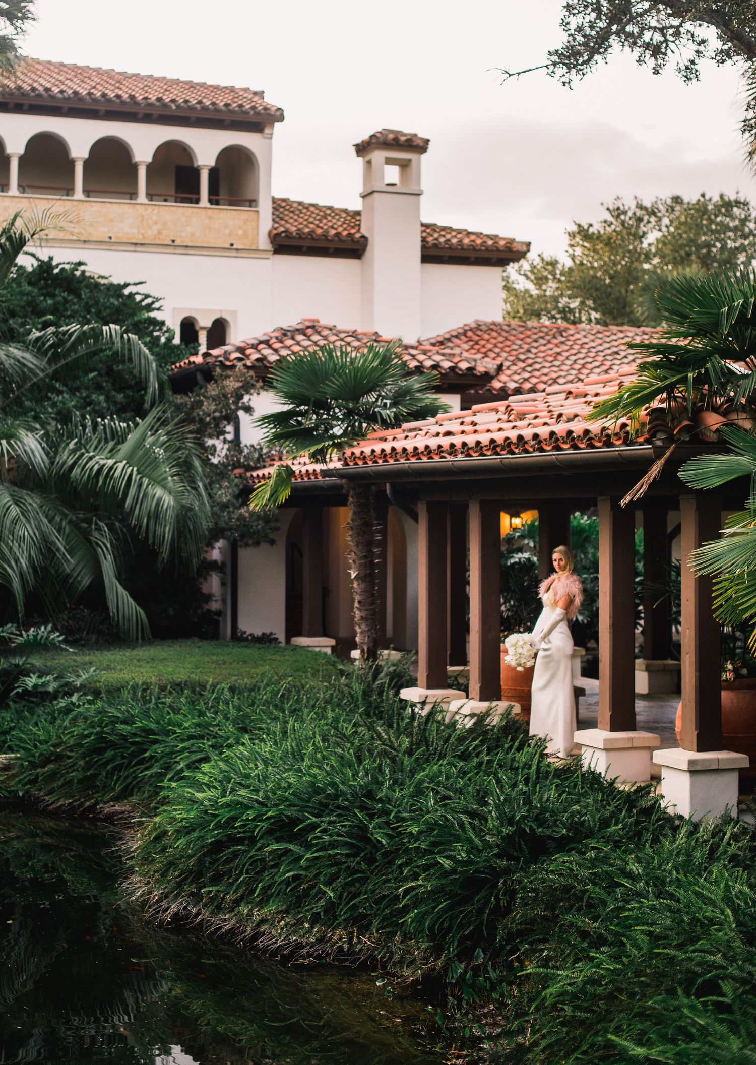 Mediterranean style building with terracotta roof tiles and white stucco walls features lush tropical landscaping and arched walkways.
