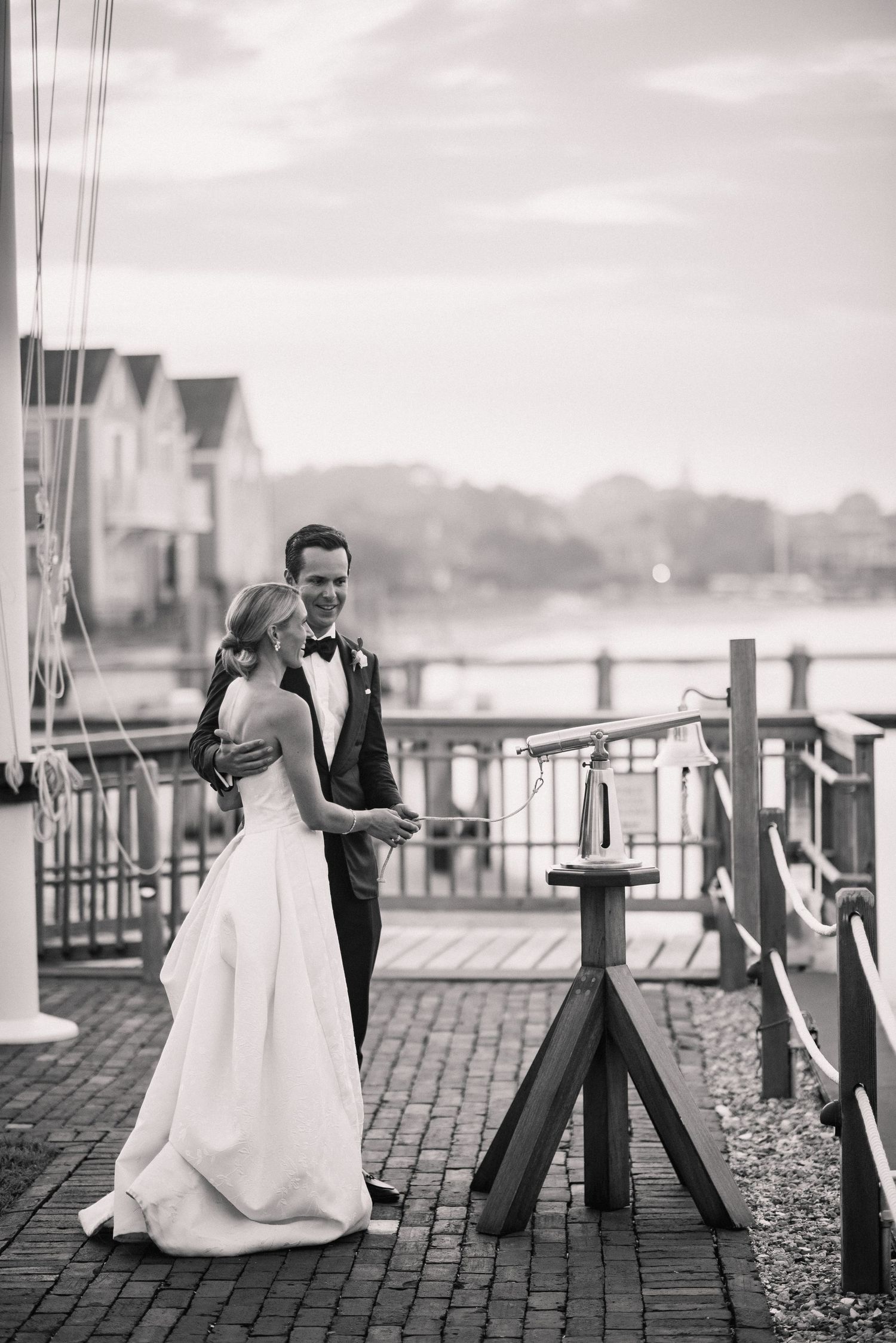 Black and white wedding portrait on a waterfront dock with urban backdrop.