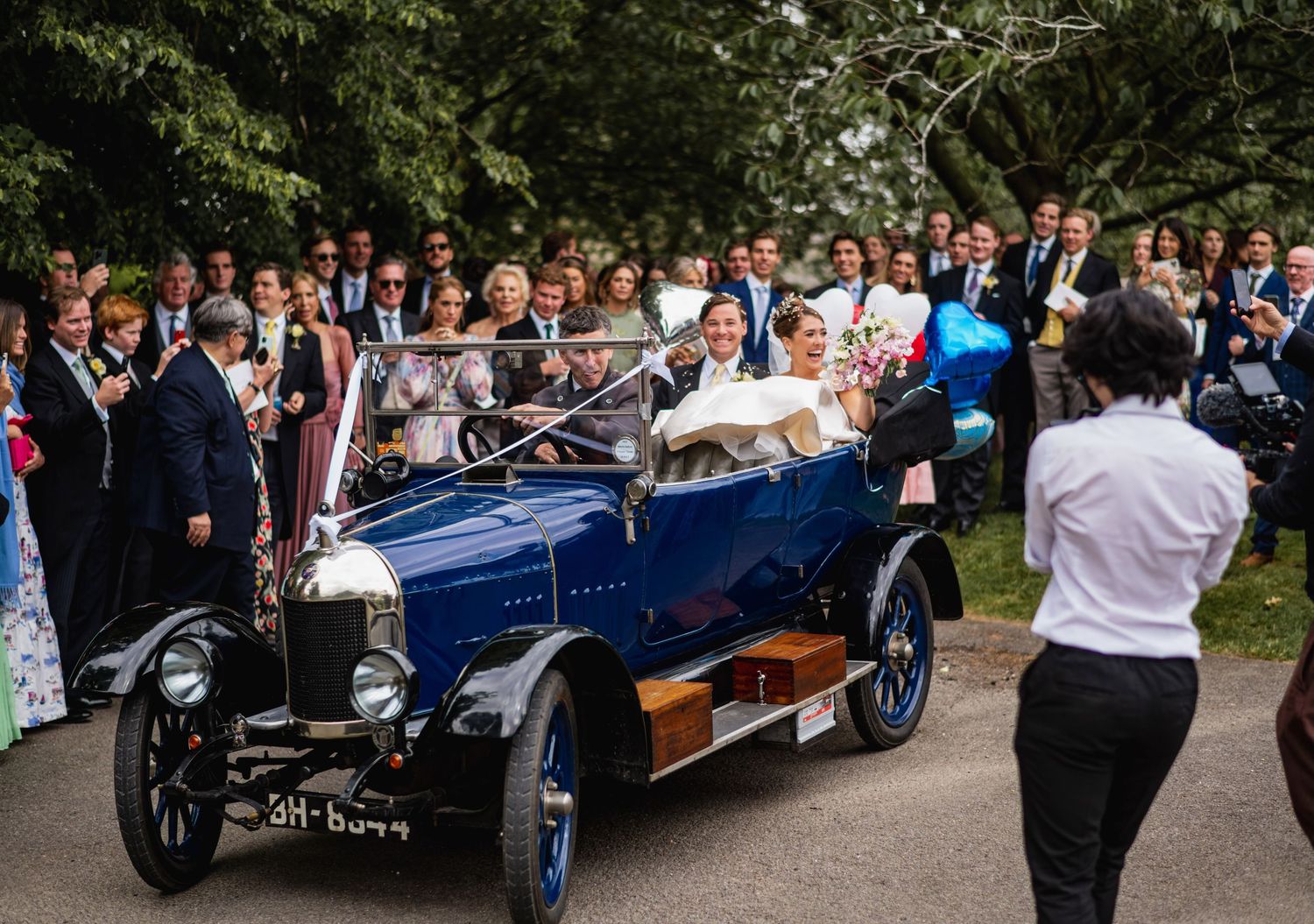 Vintage cars line up at an outdoor event with attendees gathered around on a sunny day. Cambridgeshire wedding in England