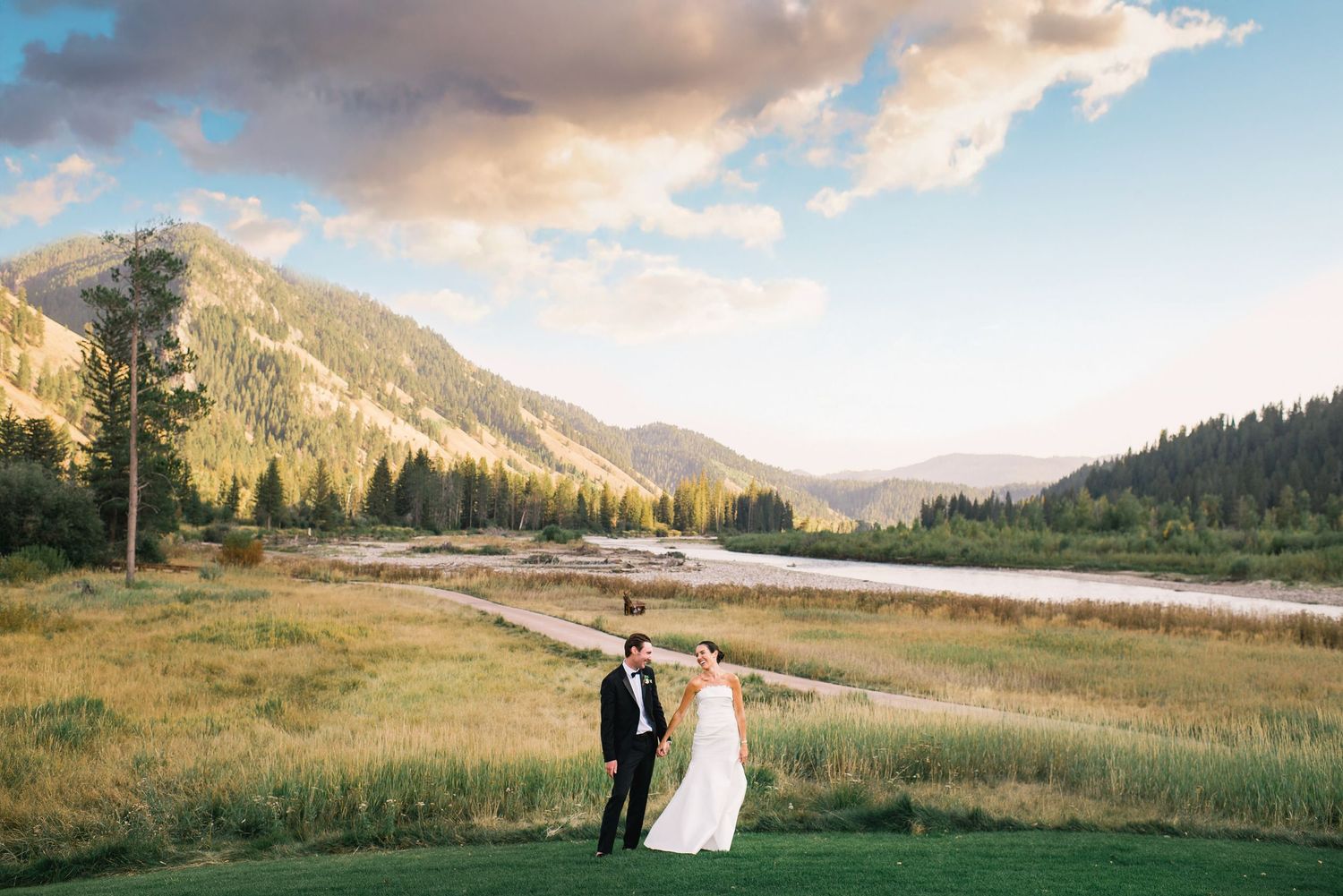 A couple stands together in a scenic mountain meadow during sunset with dramatic clouds and peaks in the background.