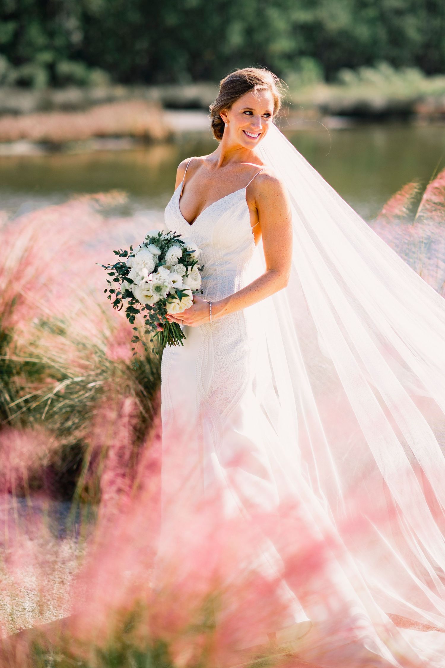 Beautiful bride in white dress holding white floral bouquet poses by lake with pink muhly grass in foreground.