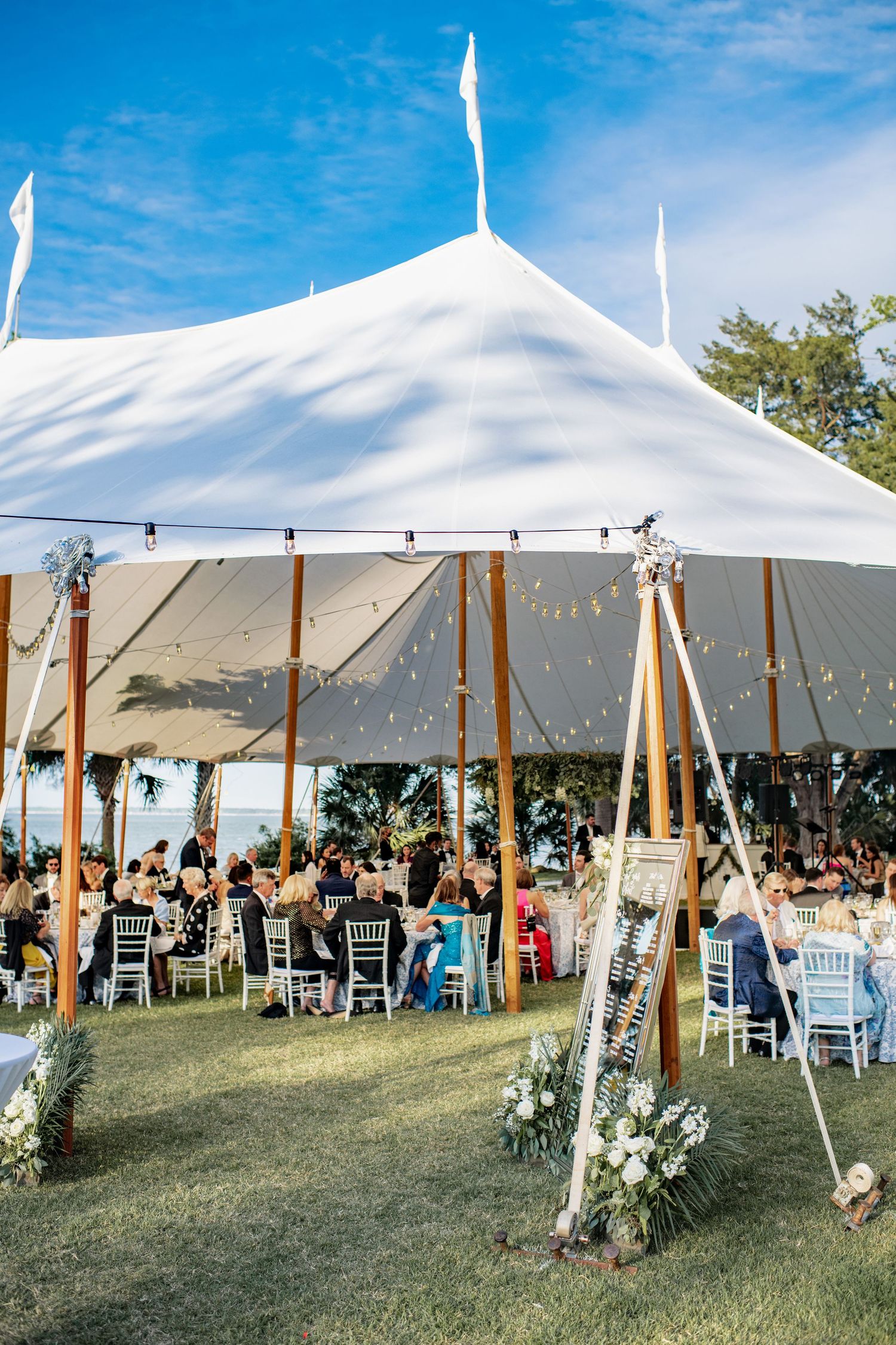 A large white wedding tent with wooden poles and floral decorations hosts guests seated at round tables under a blue sky.