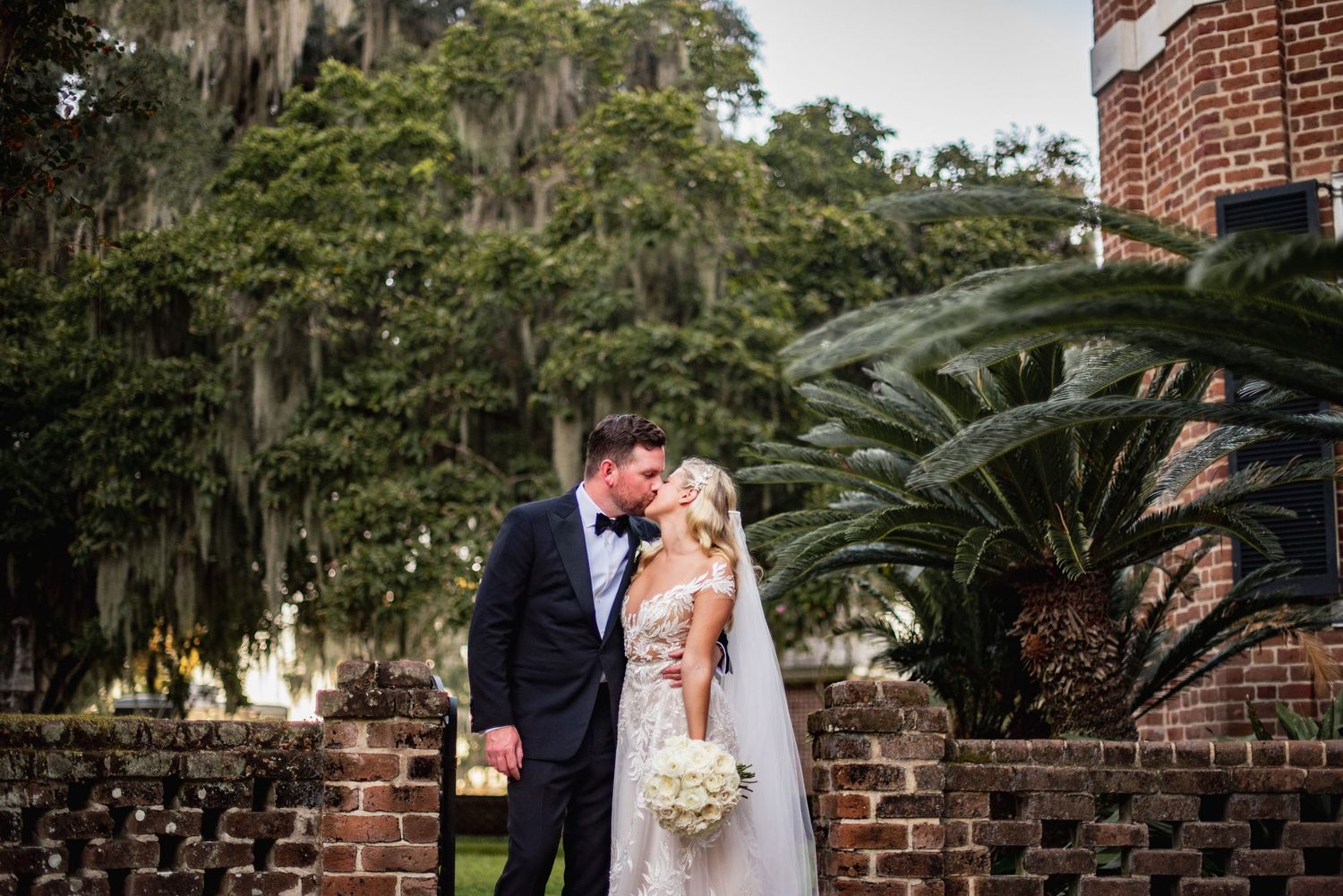 A romantic outdoor wedding ceremony takes place under Spanish moss draped oak trees and brick architecture.