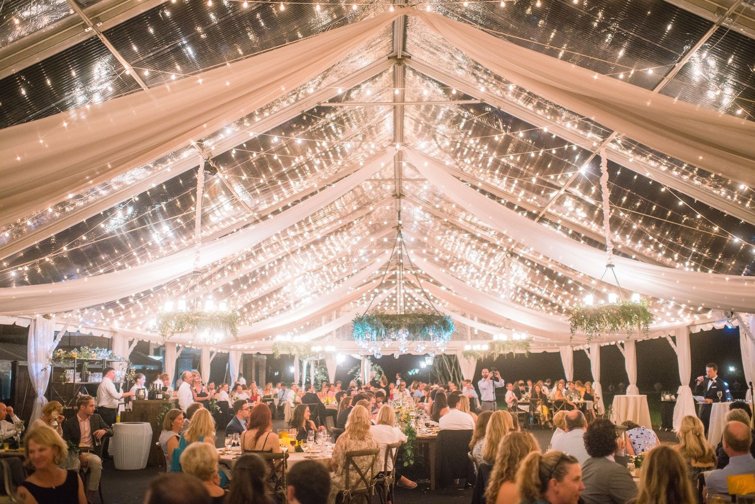 Elegant wedding reception under a clear tent decorated with string lights and white fabric draping creating a magical atmosphere.