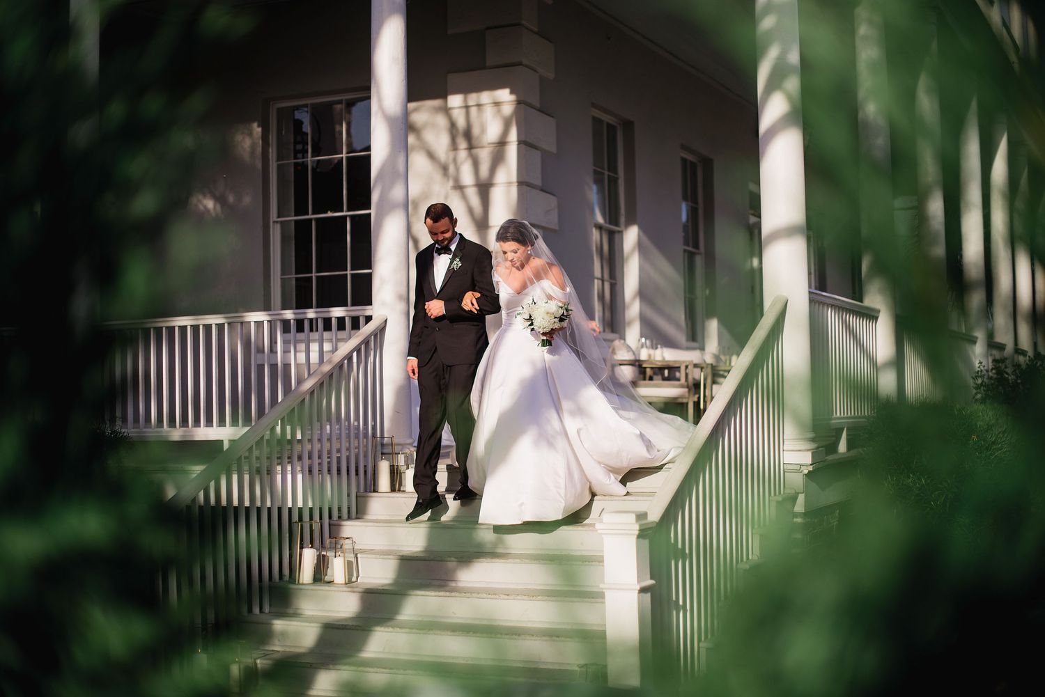 A couple in wedding attire shares a romantic moment on a white porch surrounded by lush greenery and sunlight.