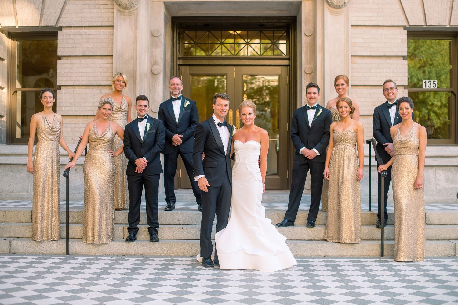 Wedding party poses in front of elegant stone columns at formal venue entrance.