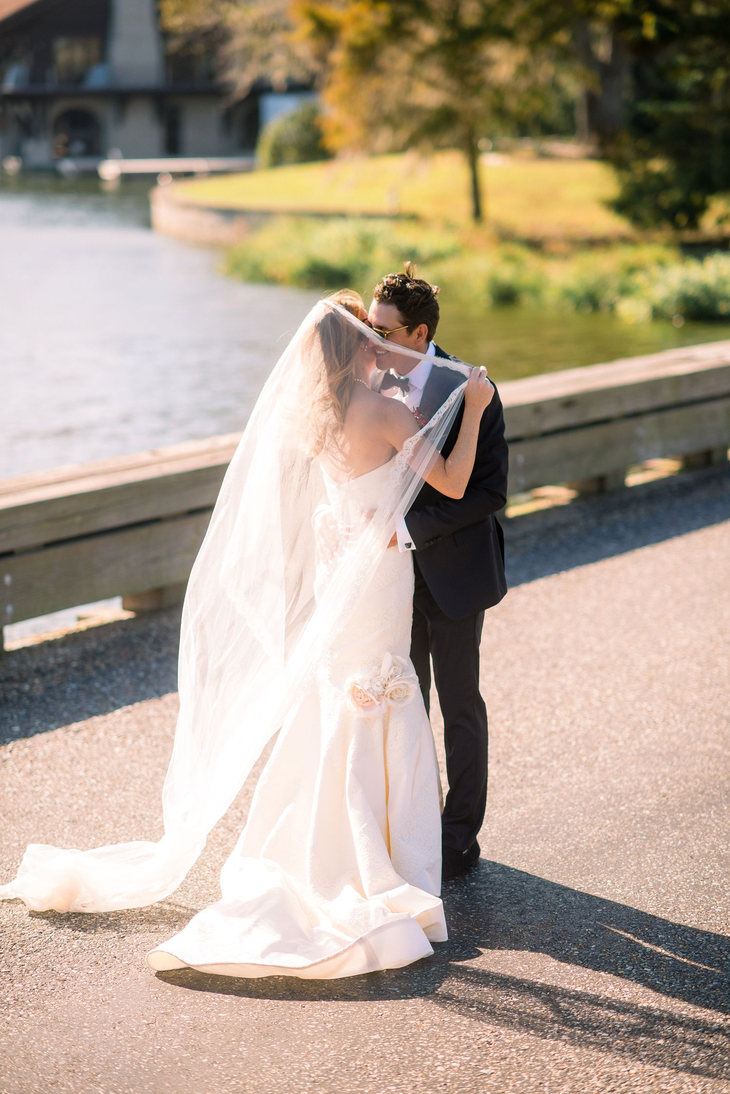 A romantic wedding couple embraces on a sunlit path, with the bride wearing a flowing white gown and long veil.