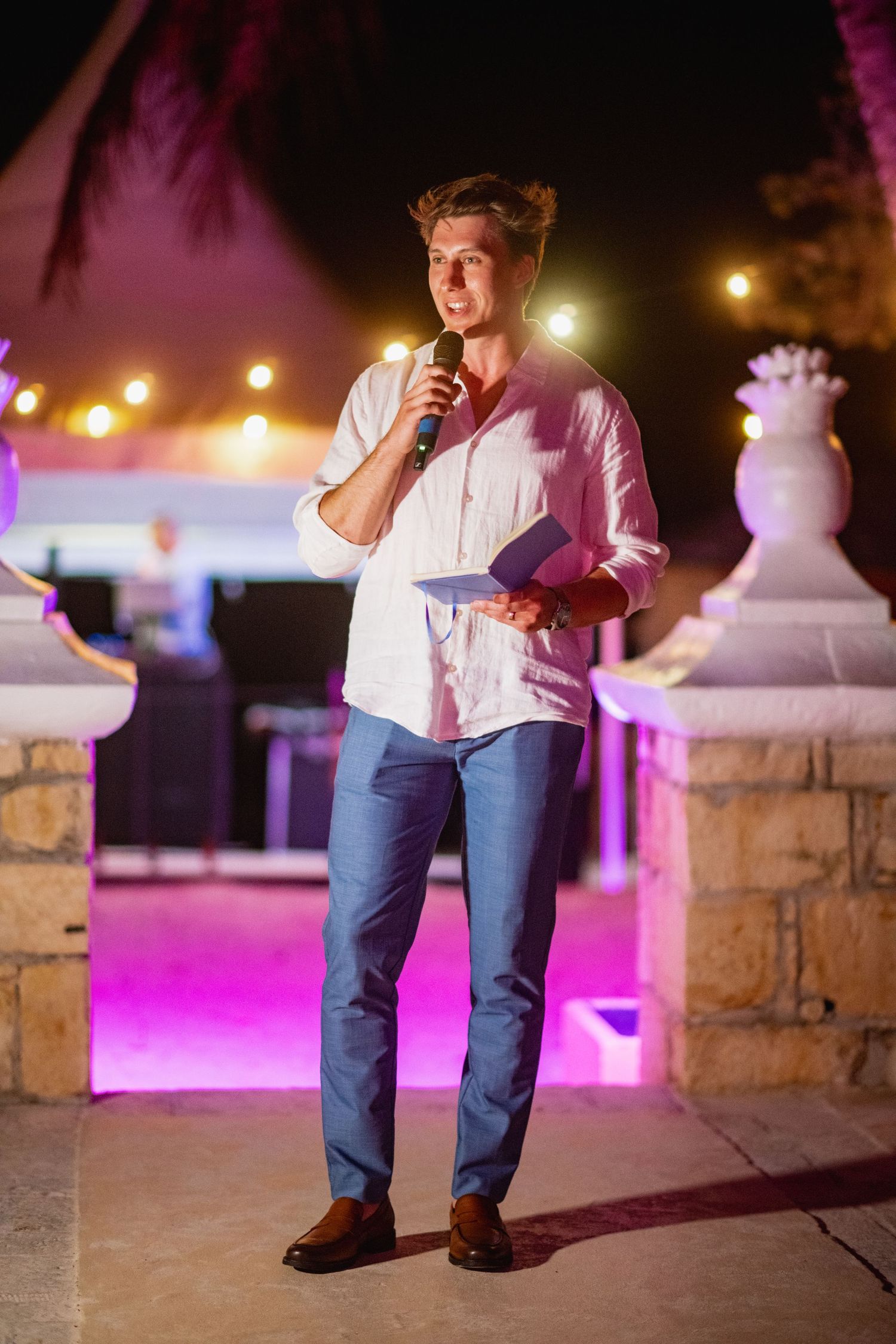 Person sitting on stone wall illuminated by pink and amber lights at night during outdoor event.