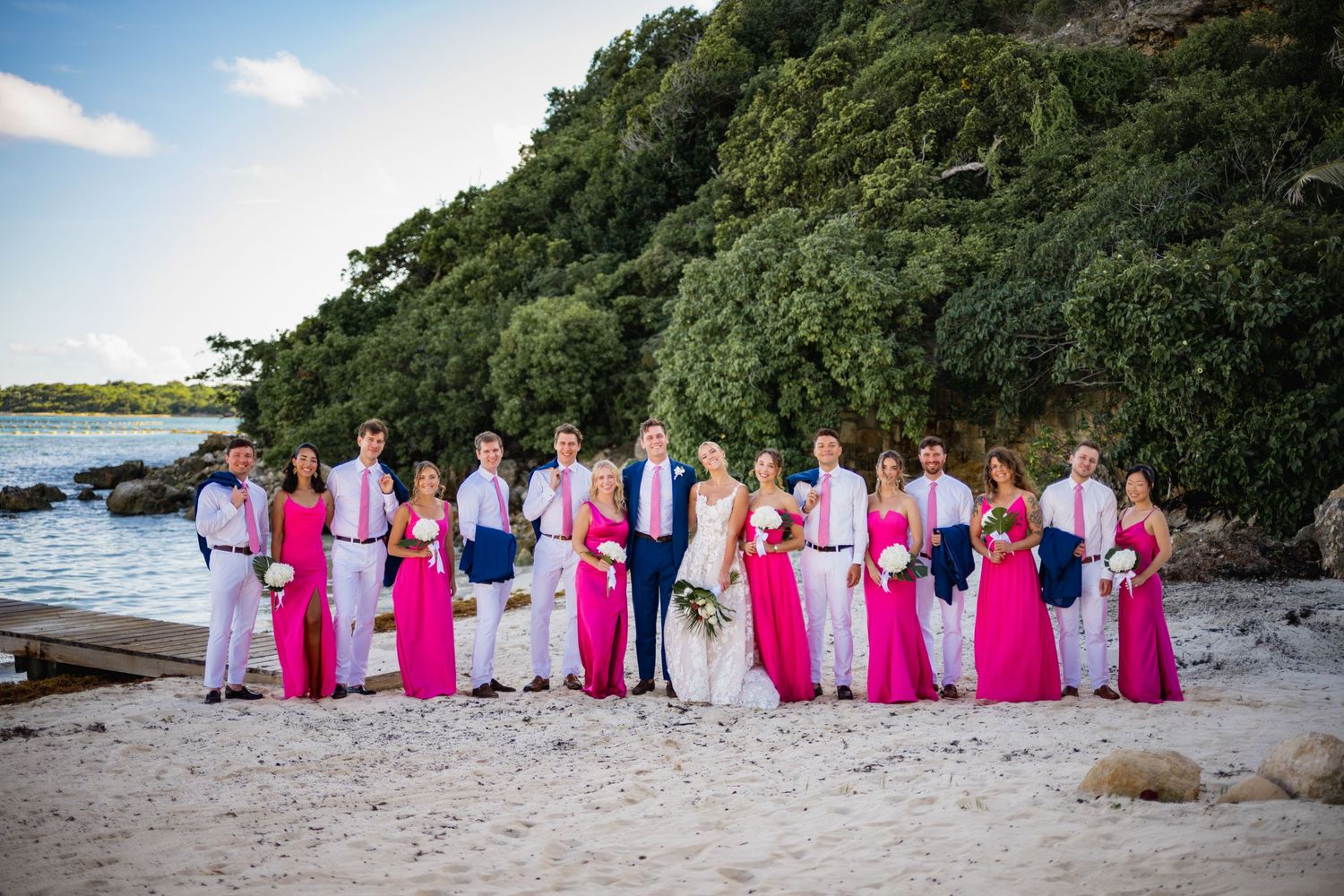Wedding party in pink and navy blue attire posed together on a tropical beach.