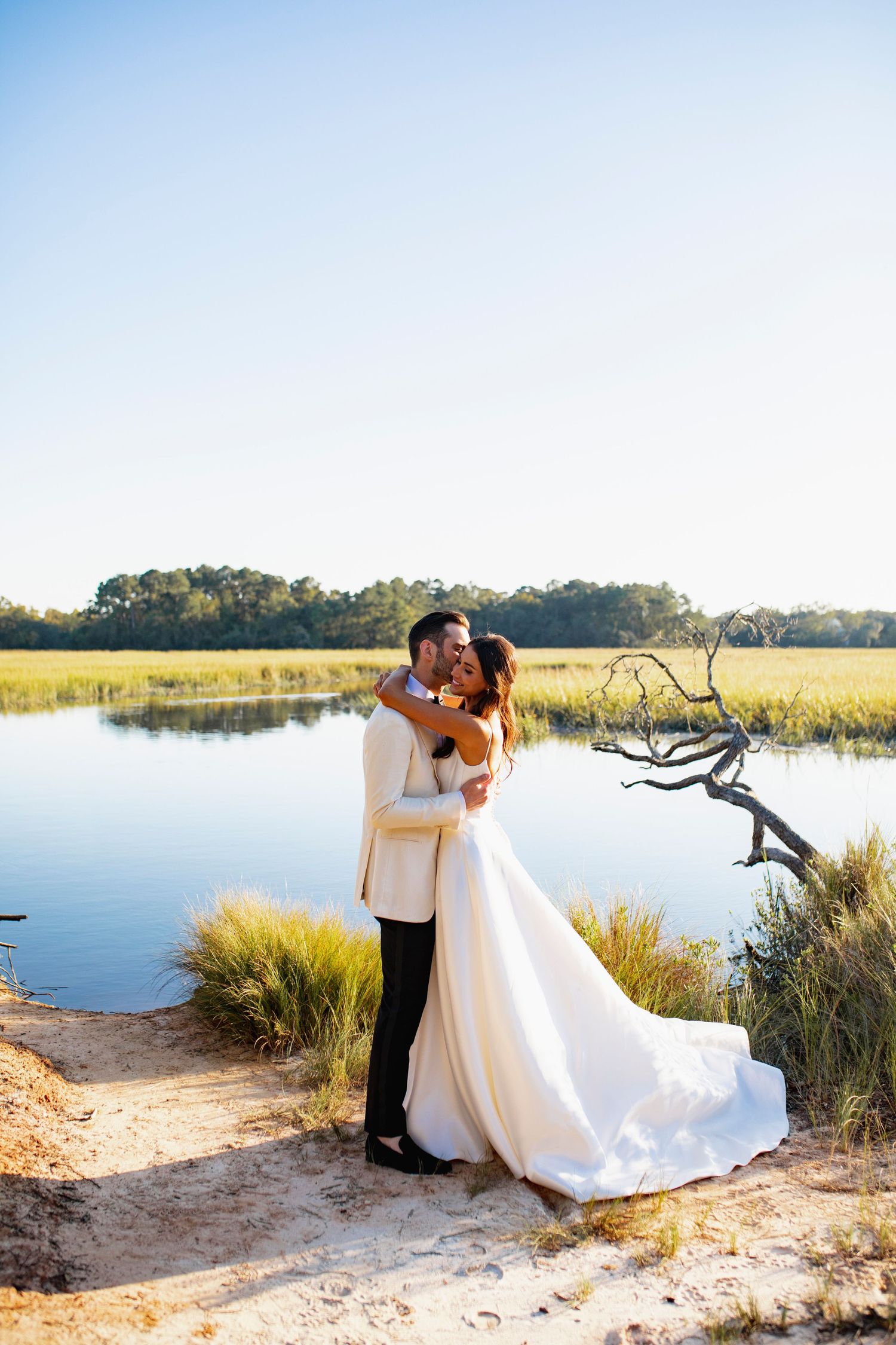 A romantic wedding photoshoot at sunset by a serene lake with tall grasses and natural scenery in the background.