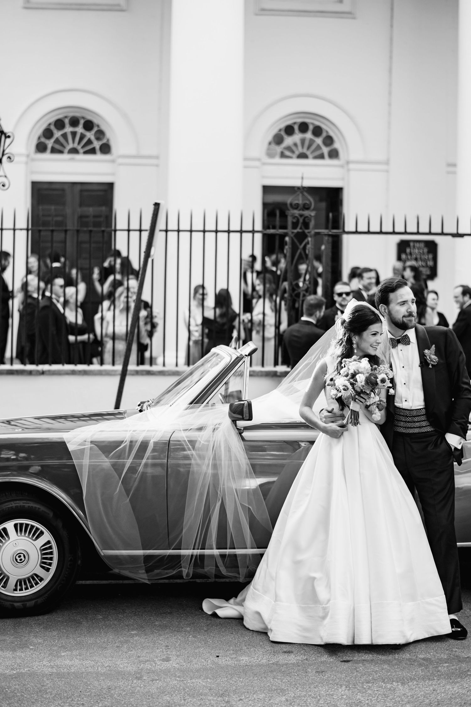 A black and white wedding photo series showing a couple posing with a classic car outside an elegant white building.