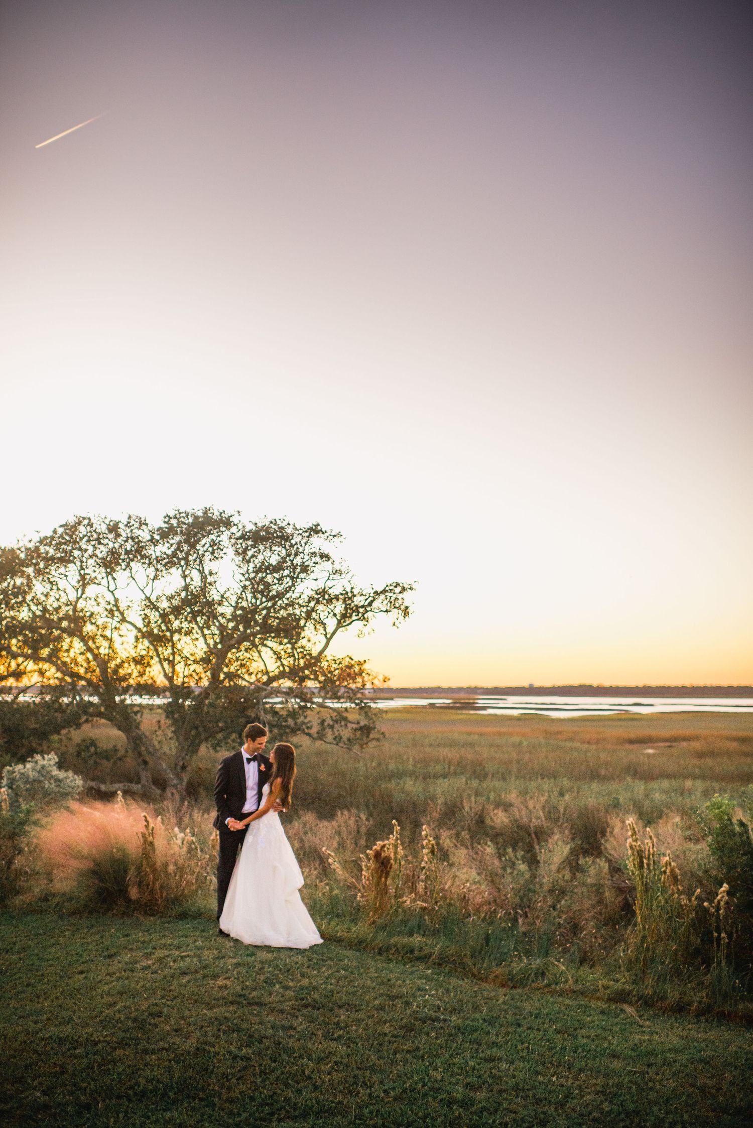 Romantic wedding couple silhouetted against a golden sunset over coastal marshlands during their outdoor photo session.