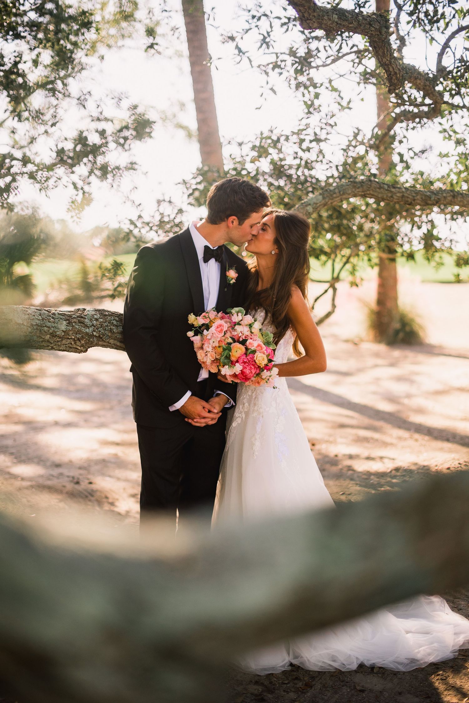 A couple in wedding attire shares an intimate moment outdoors under sunlit trees in a romantic garden setting.