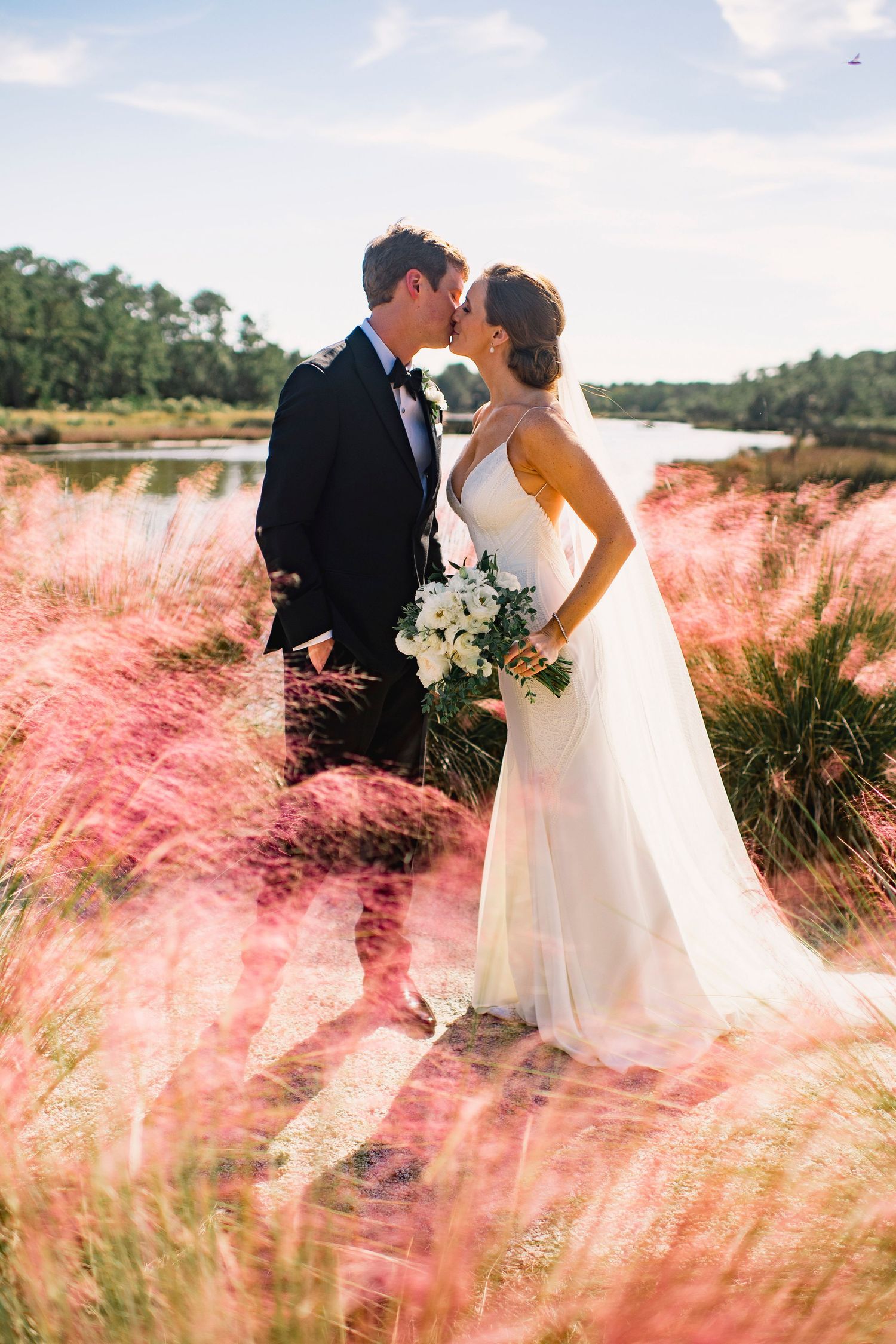Romantic wedding couple embraces in field of pink muhly grass during golden hour by lakeside.